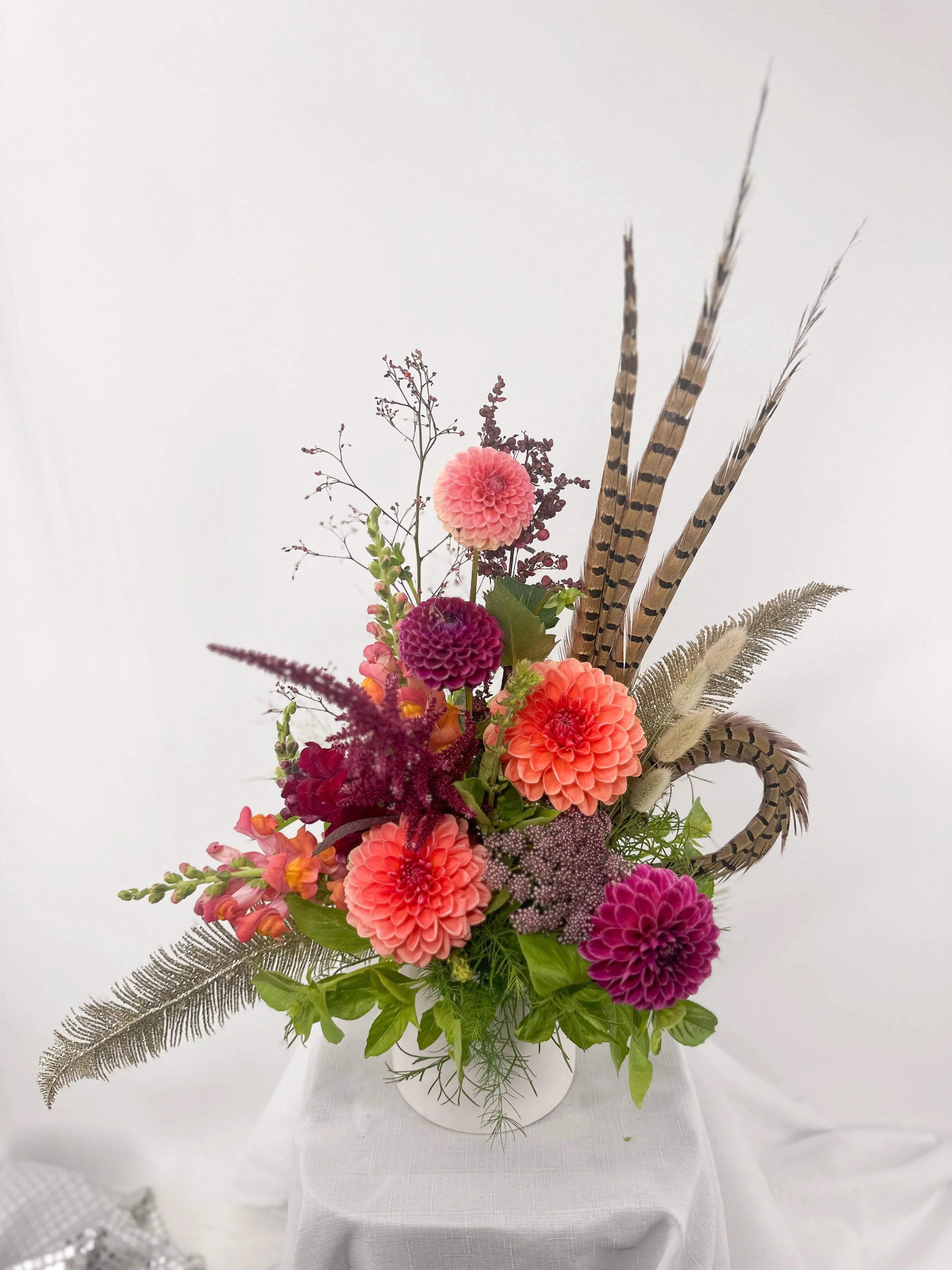 Colorful flower arrangement with pink, purple, peach, and red dahlias, fern leaves, and decorative feathers in a white vase on a white table.