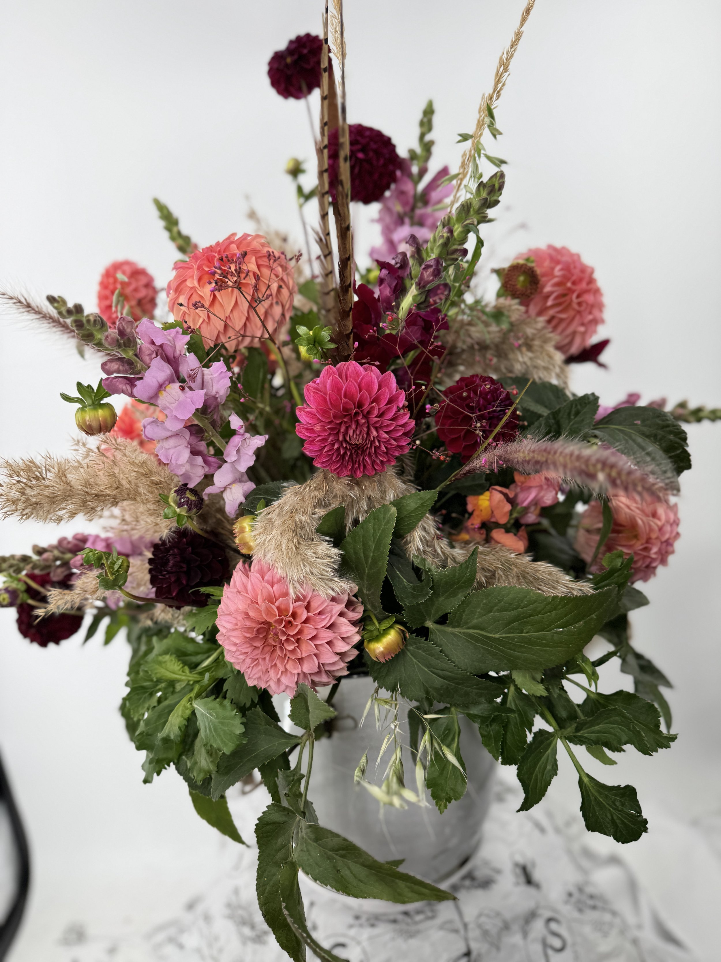 Colorful mixed flower bouquet in a gray vase, featuring pink, purple, and deep red blooms, with greenery and dried grasses.