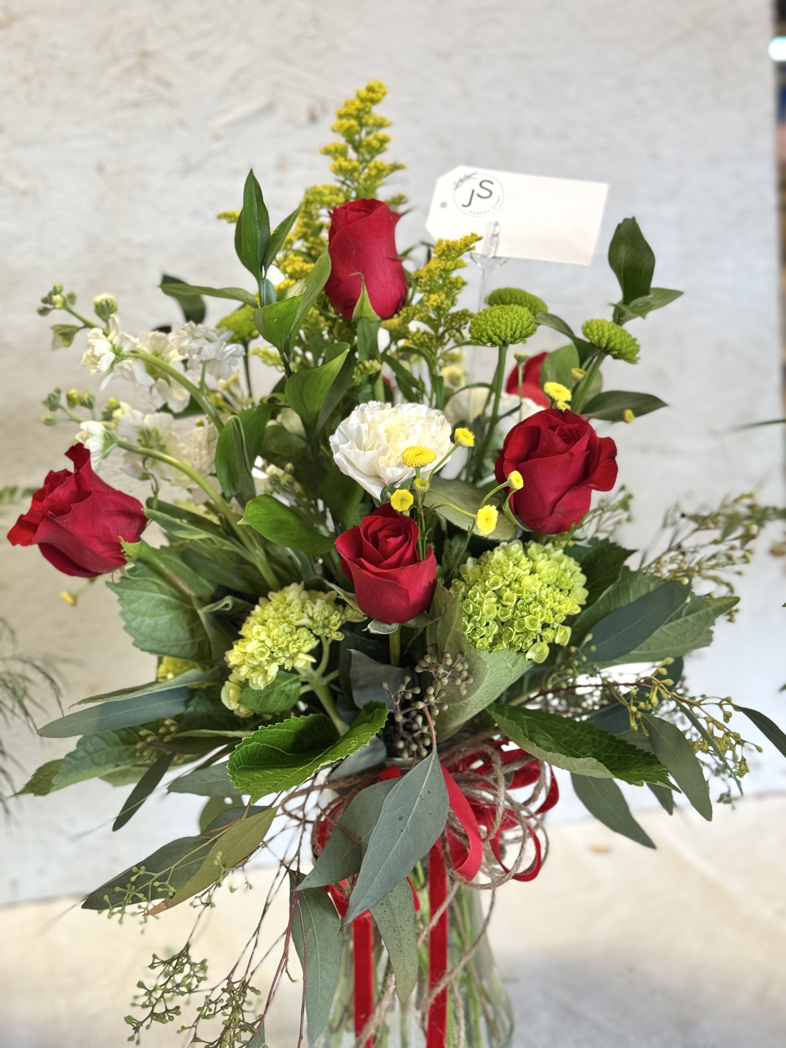 A floral arrangement with red roses, white carnations, green hydrangeas, and yellow filler flowers, tied with a red ribbon.