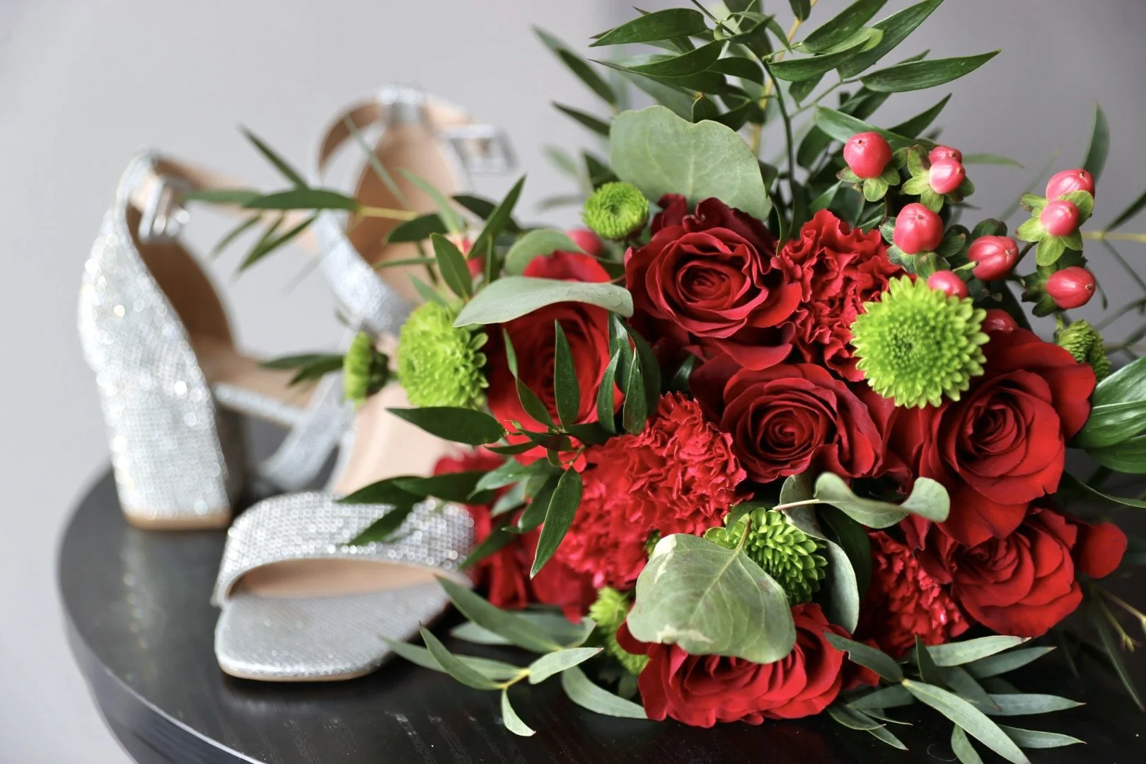 A bouquet of red roses, carnations, and green chrysanthemums with pink berries and green leaves, accompanied by a pair of silver high-heeled shoes with rhinestone embellishments, placed on a black surface.