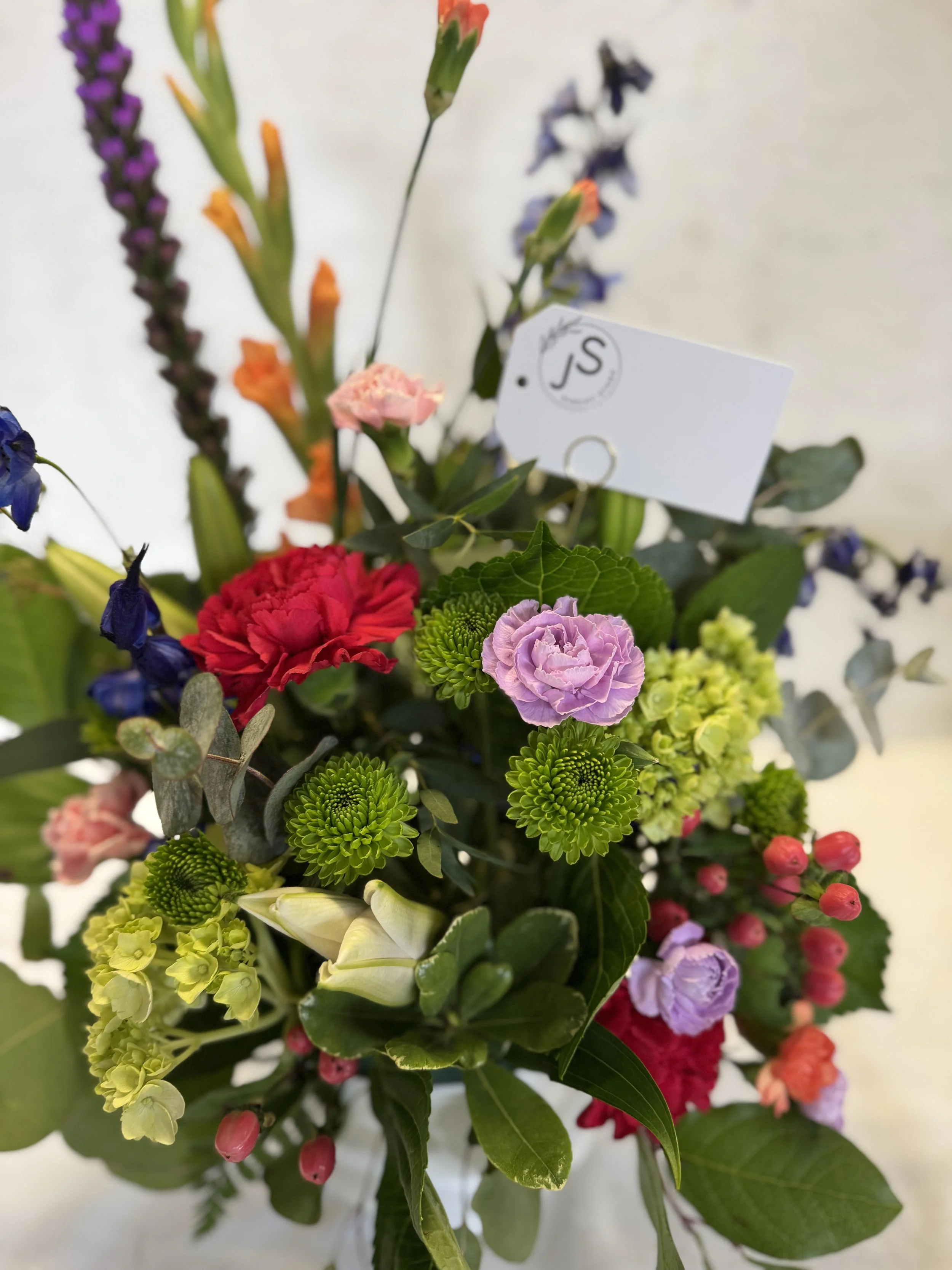 Colorful flower bouquet with pink, purple, red, yellow, and orange flowers, and green leaves, with a white tag attached.
