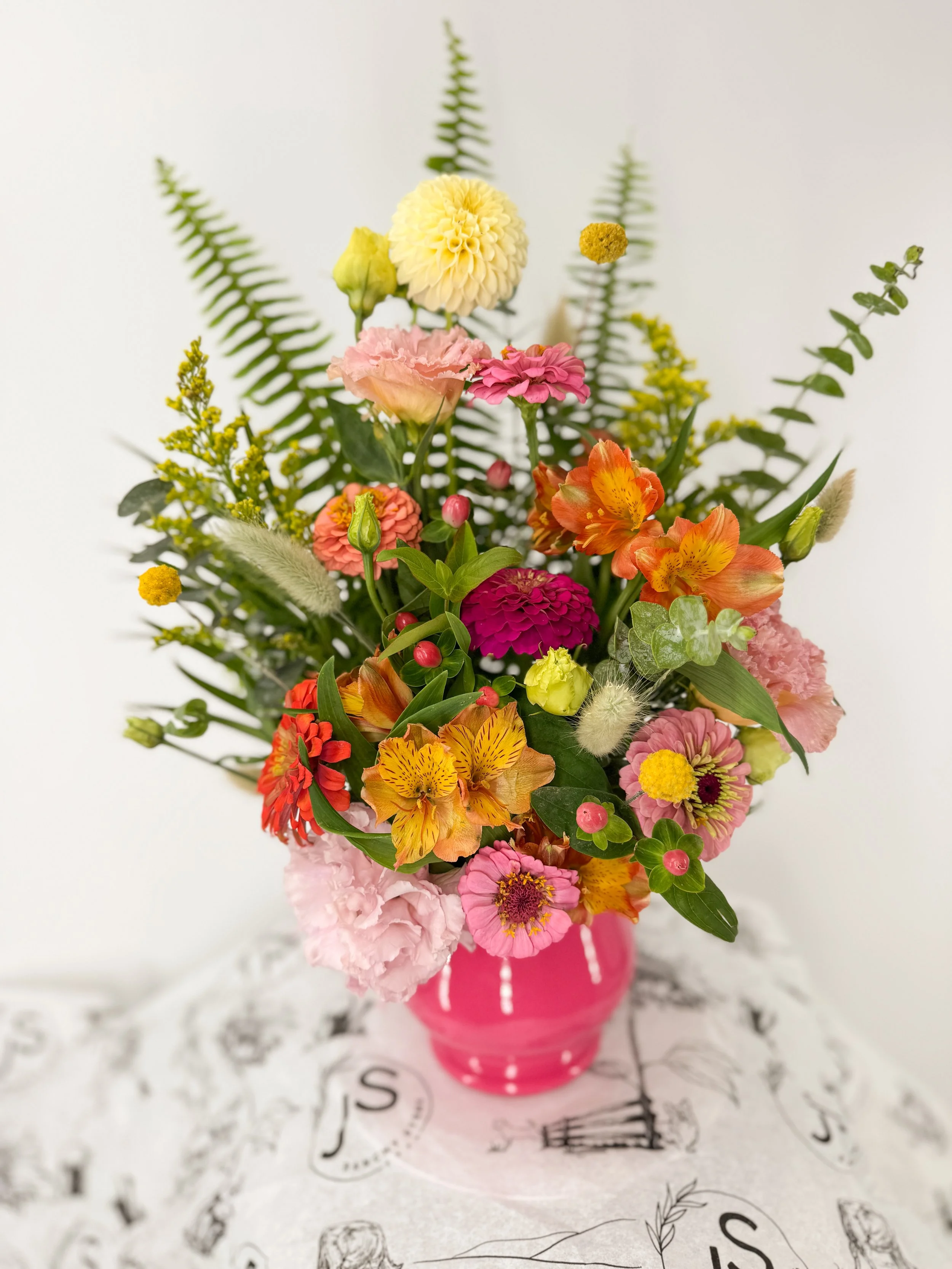 A colorful flower arrangement in a pink vase, featuring pink, orange, yellow, and white blooms surrounded by green fern leaves and other greenery.
