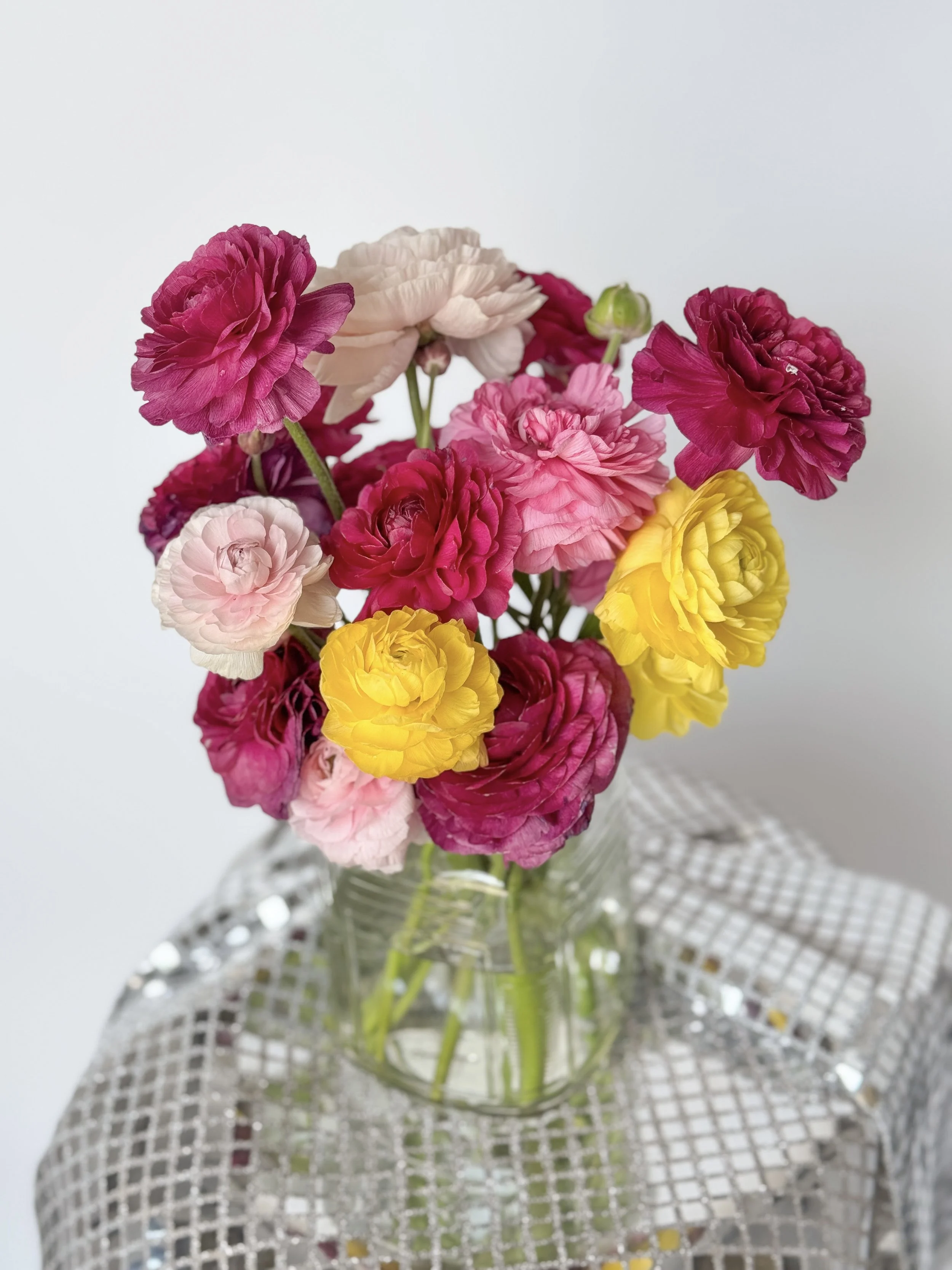 A bouquet of colorful ranunculus flowers in a clear glass vase on a silver tablecloth.