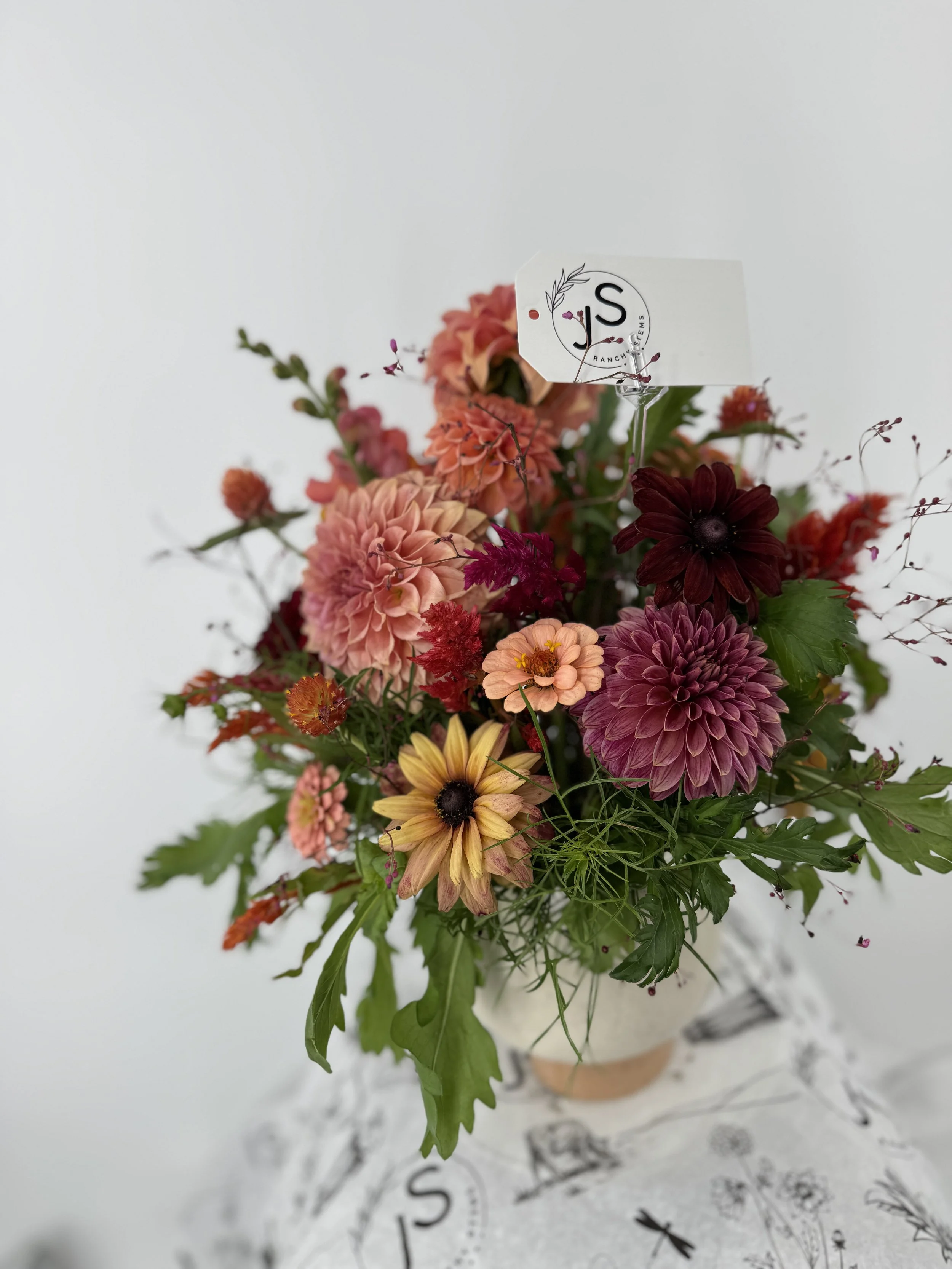 A colorful flower bouquet with various pink, red, yellow, and purple blossoms arranged in a vase on a table with botanical print.