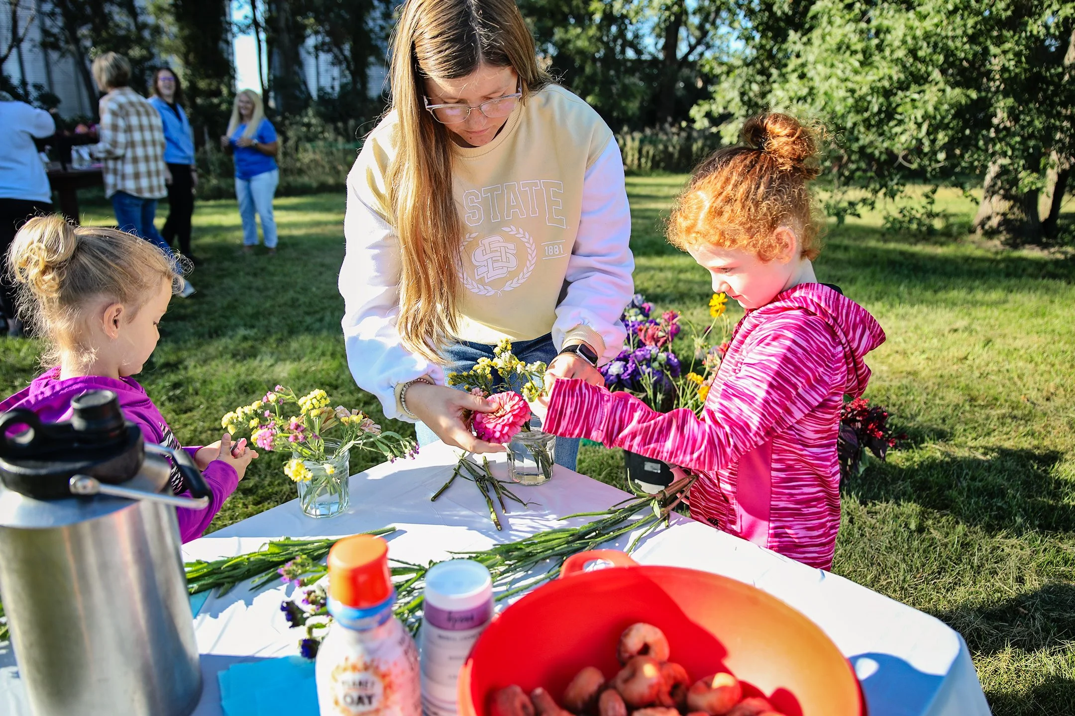 A woman and two young girls arranging flowers on a table outdoors during daytime, with several people in the background.