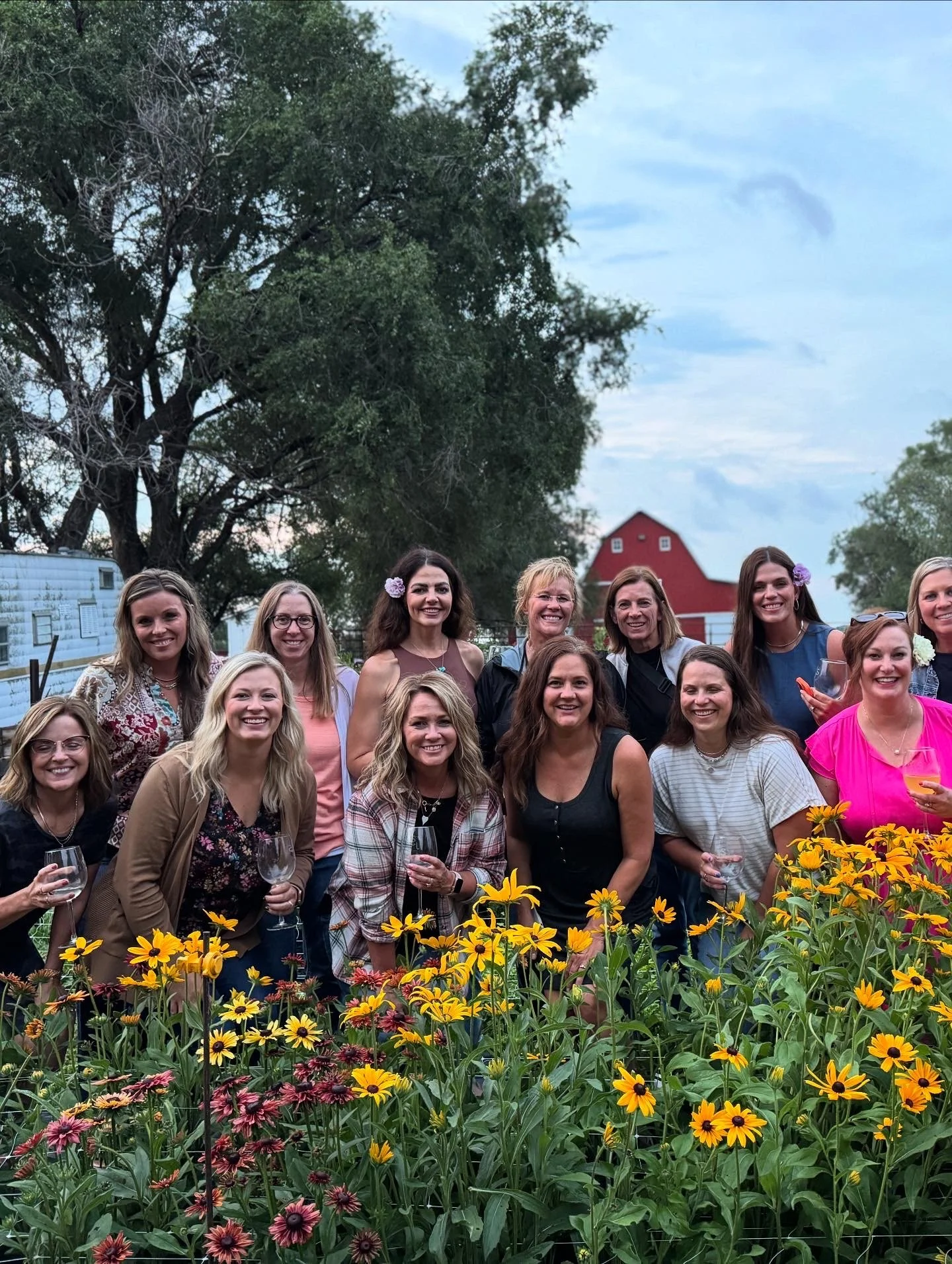 A group of women smiling and holding wine glasses outdoors in front of a flower garden with a barn and trees in the background.