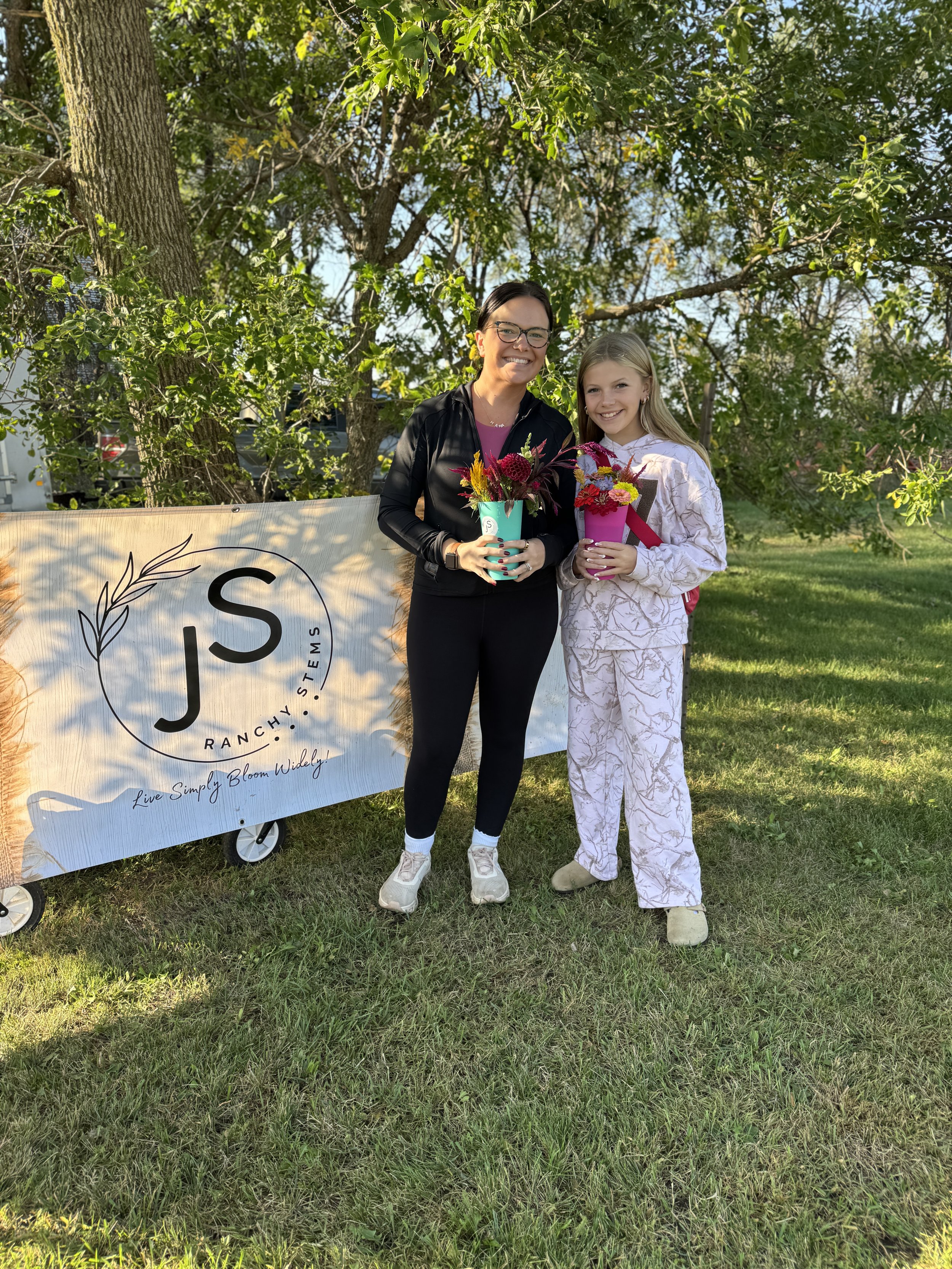 A woman and girl standing on grass outdoors, holding colorful flower bouquets, smiling in front of a tree and a sign with the text 'J S Ranchy. Stem. Live Simply. Bloom Widely.'