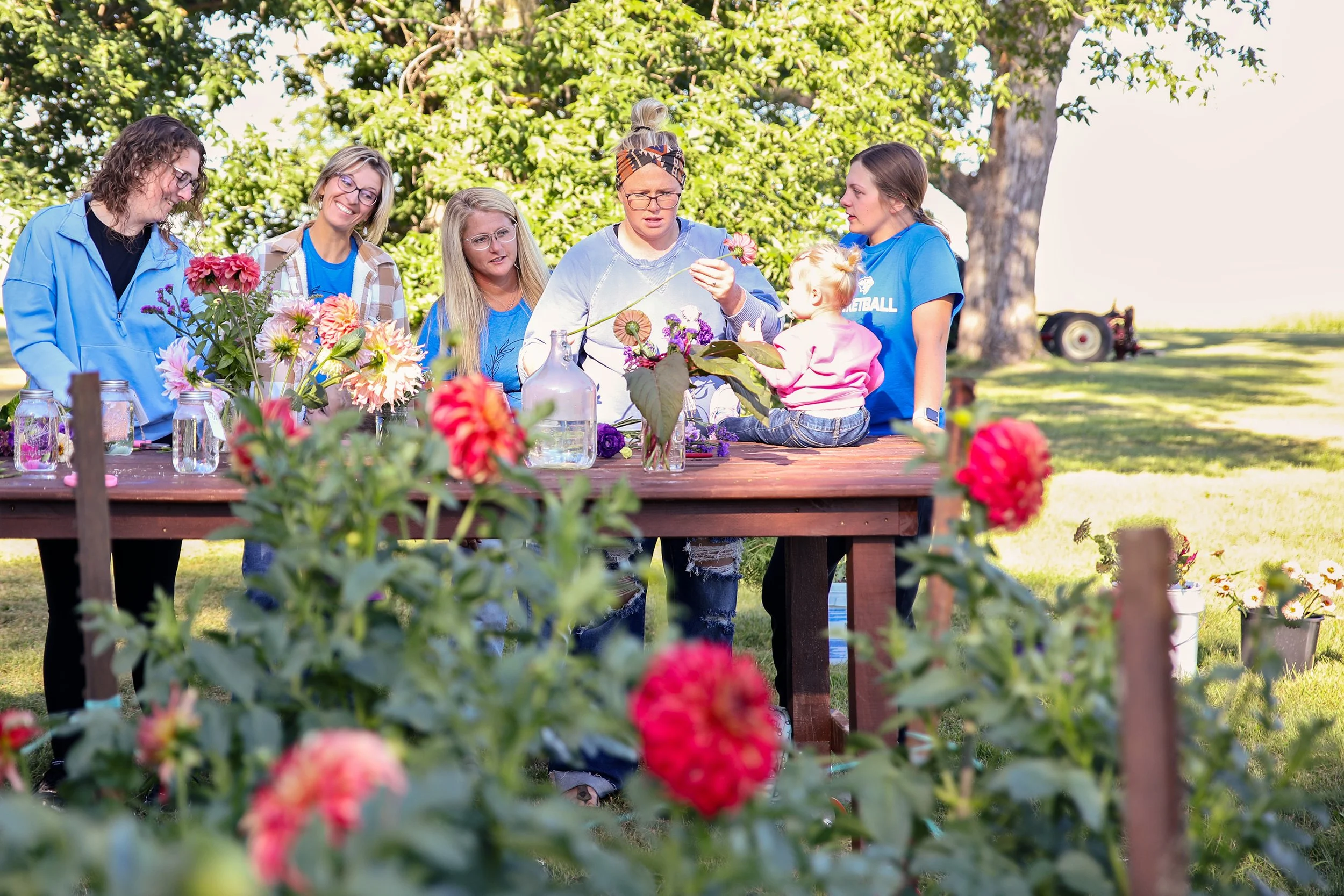 A group of six women and one young girl are gathered around a wooden table outdoors, arranging and admiring flowers in vases, with trees and sunshine in the background.