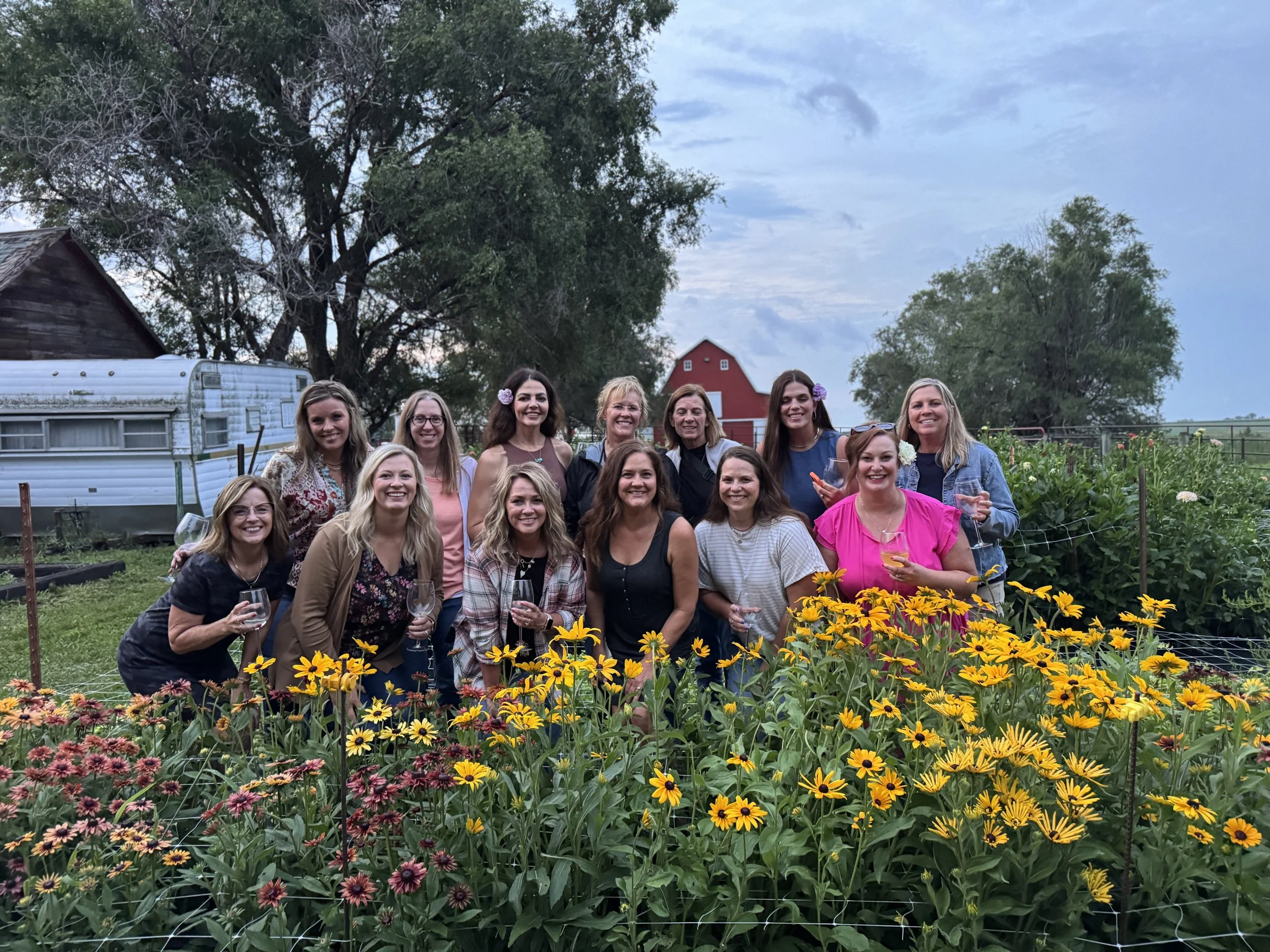 A group of fifteen women gathered in a garden with yellow and pink flowers, holding glasses of wine, smiling at the camera.