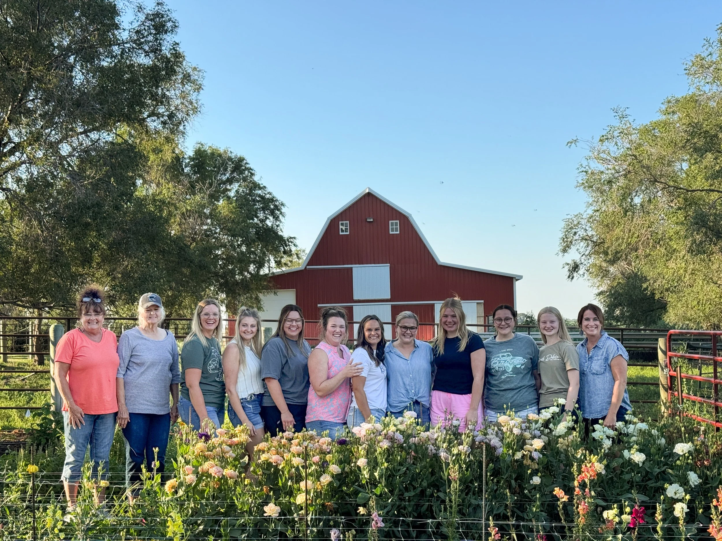 A group of women standing in front of a red barn and flowering plants on a farm, with trees and blue sky in the background.