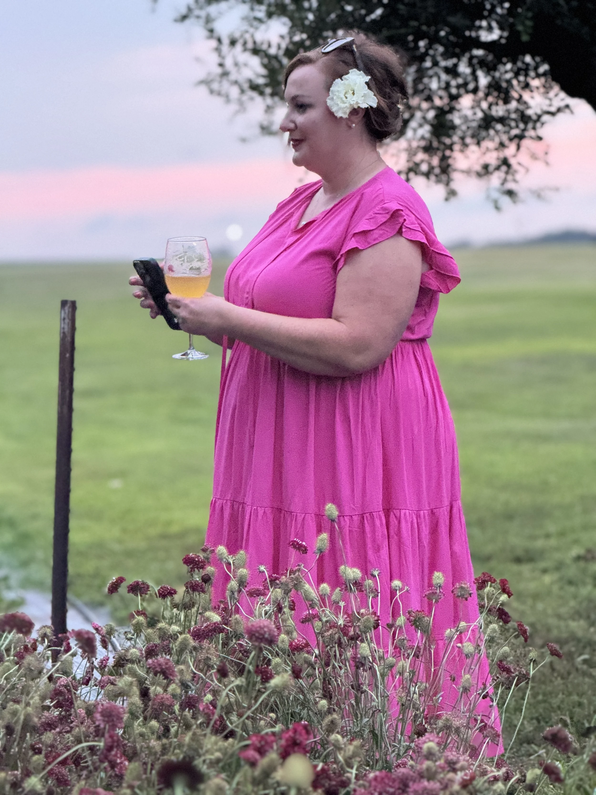 A woman in a pink dress with a white flower in her hair holding a glass of beer and a phone, standing outdoors in a field with pink flowers and a sunset in the background.