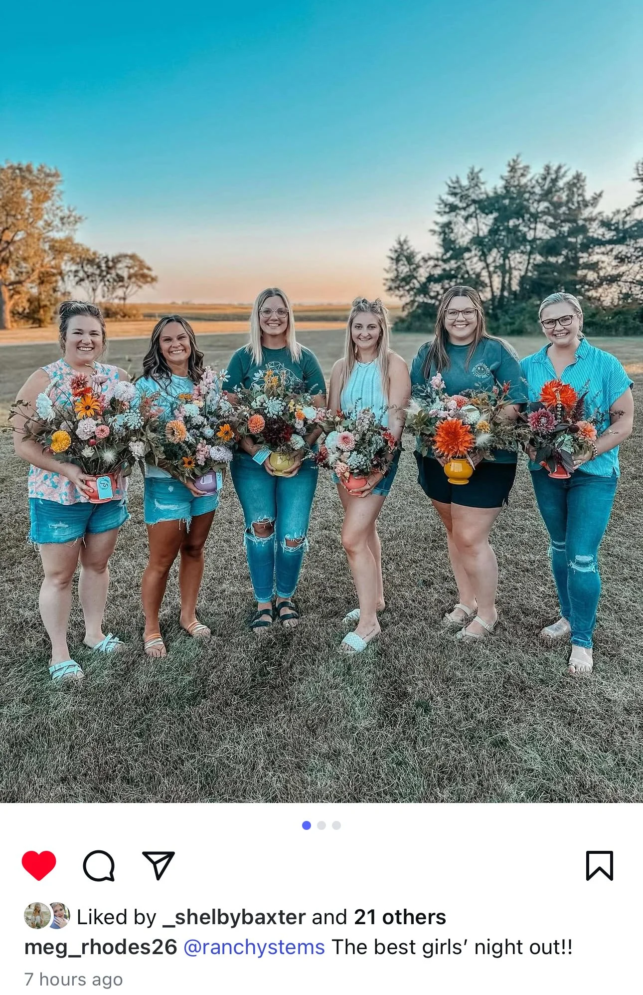 Six women standing outdoors on grass at sunset, each holding a bouquet of colorful flowers, smiling at the camera.