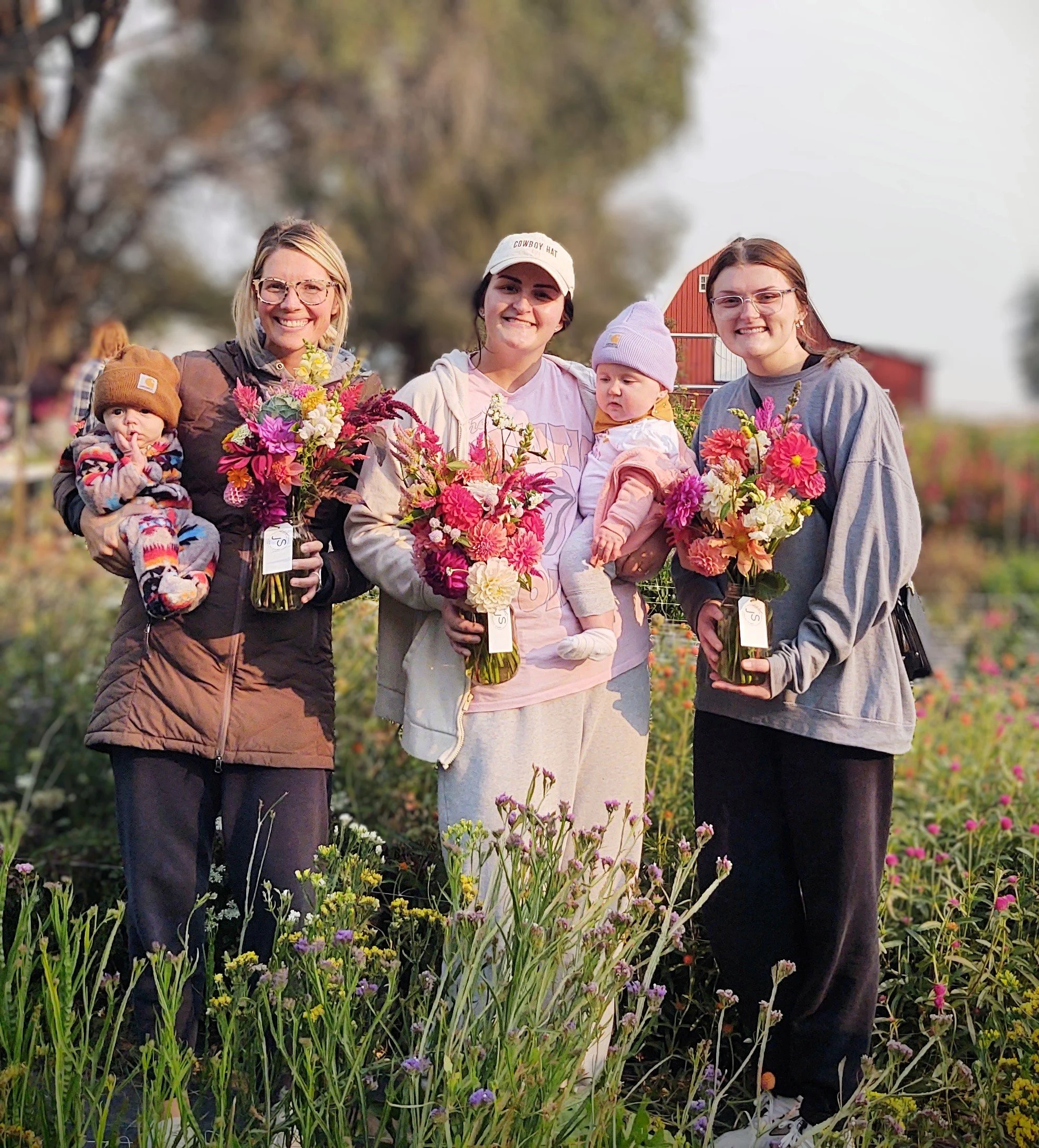 Four women and two children standing outdoors in a flower field, holding colorful flower bouquets, smiling at the camera.
