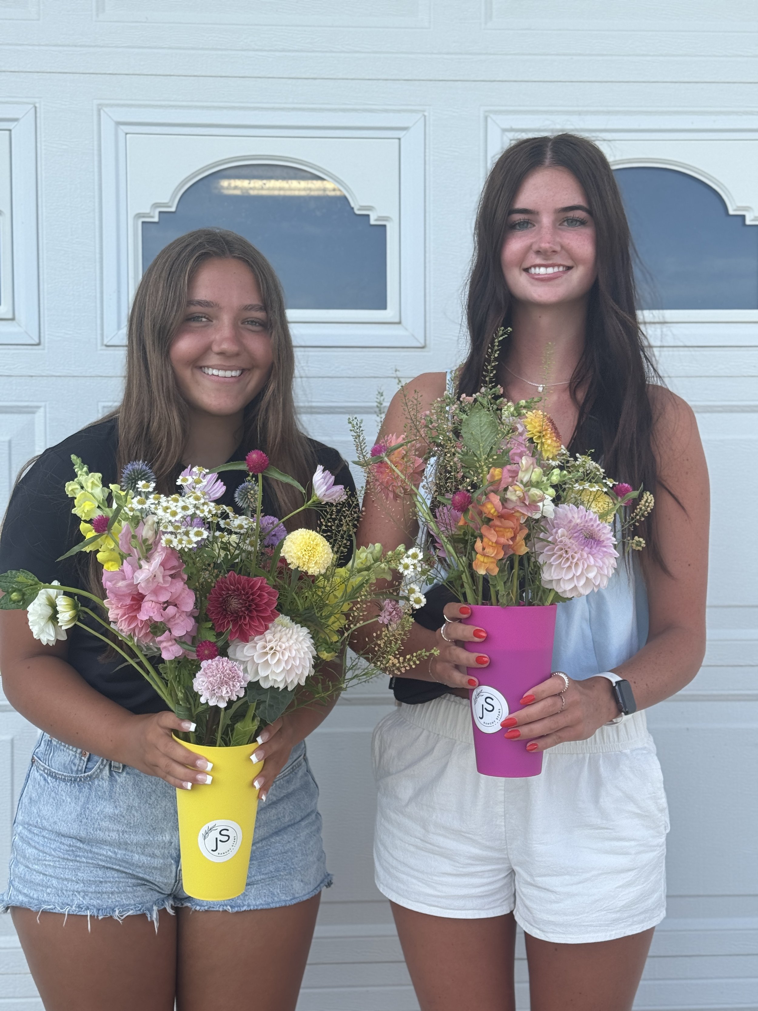 Two young women standing in front of a white garage door, each holding colorful flower arrangements in vases.