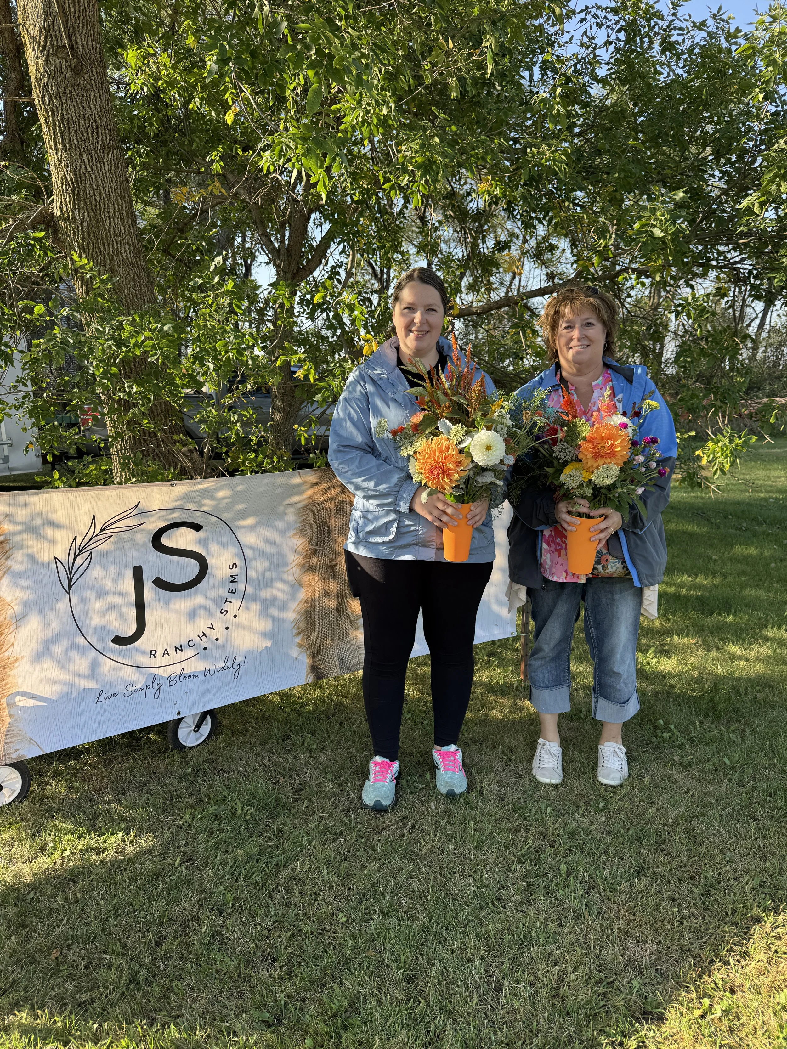 Two women standing outdoors in front of a sign that reads "J S Ranchy. Systems, Live Simply Bloom Widely!" Each woman holding a colorful bouquet of flowers, smiling at the camera.