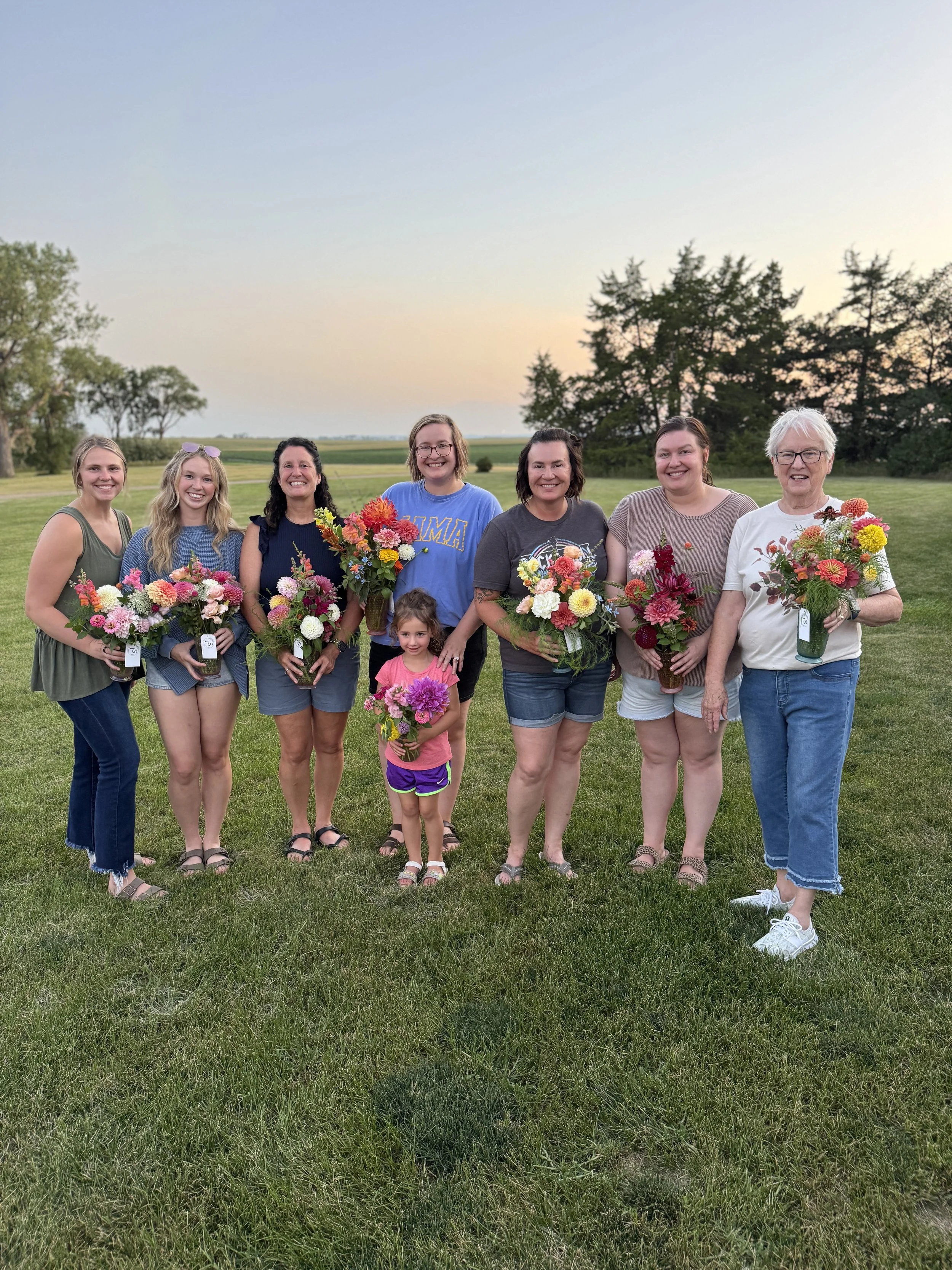 Nine people, including children, standing outdoors on grass, holding colorful flower bouquets, during sunset or early evening with trees and open field in the background.