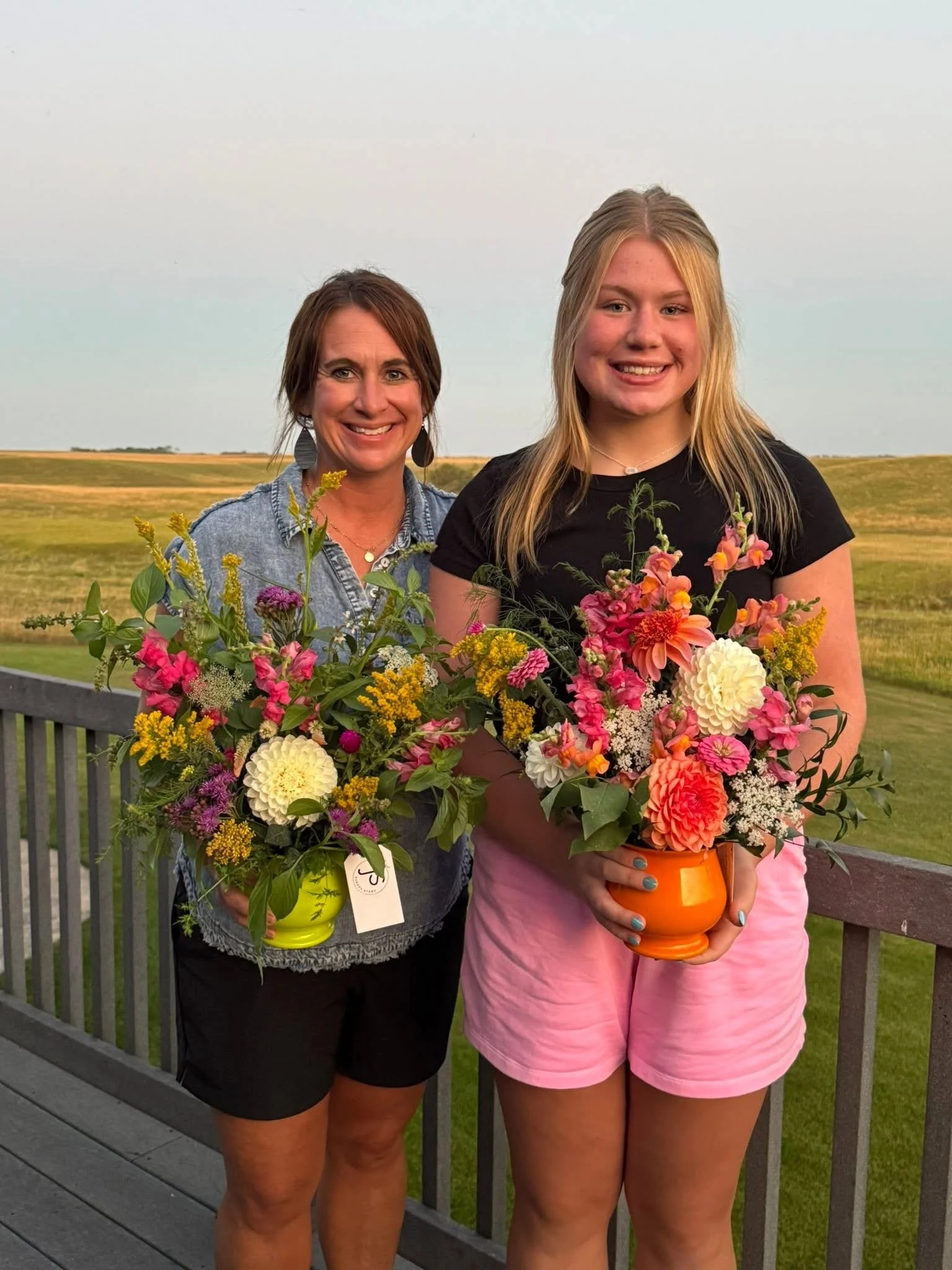Two women standing outdoors on a wooden deck holding colorful flower arrangements in pots, with a rural landscape in the background.