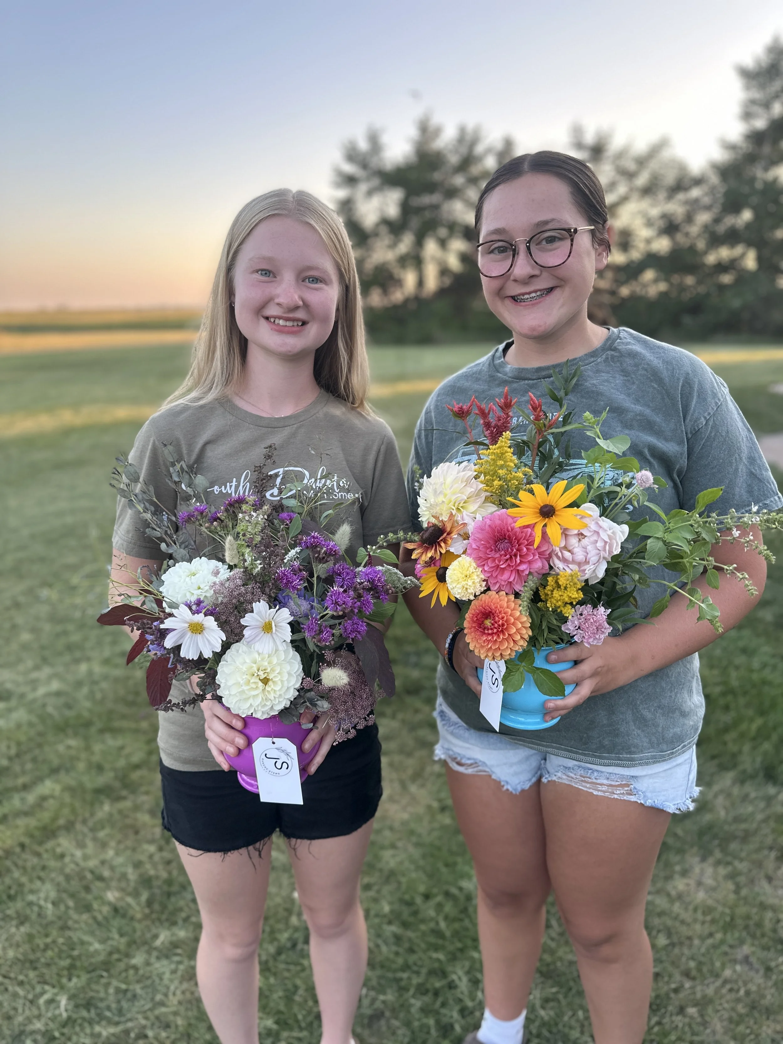 Two young girls outdoors at sunset, holding colorful flower arrangements in small buckets, smiling at the camera.
