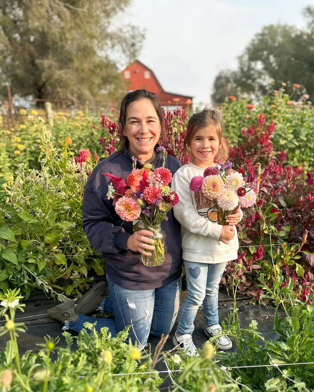 Two smiling girls holding colorful flower bouquets in a garden with blooming flowers and greenery, and a red barn in the background.