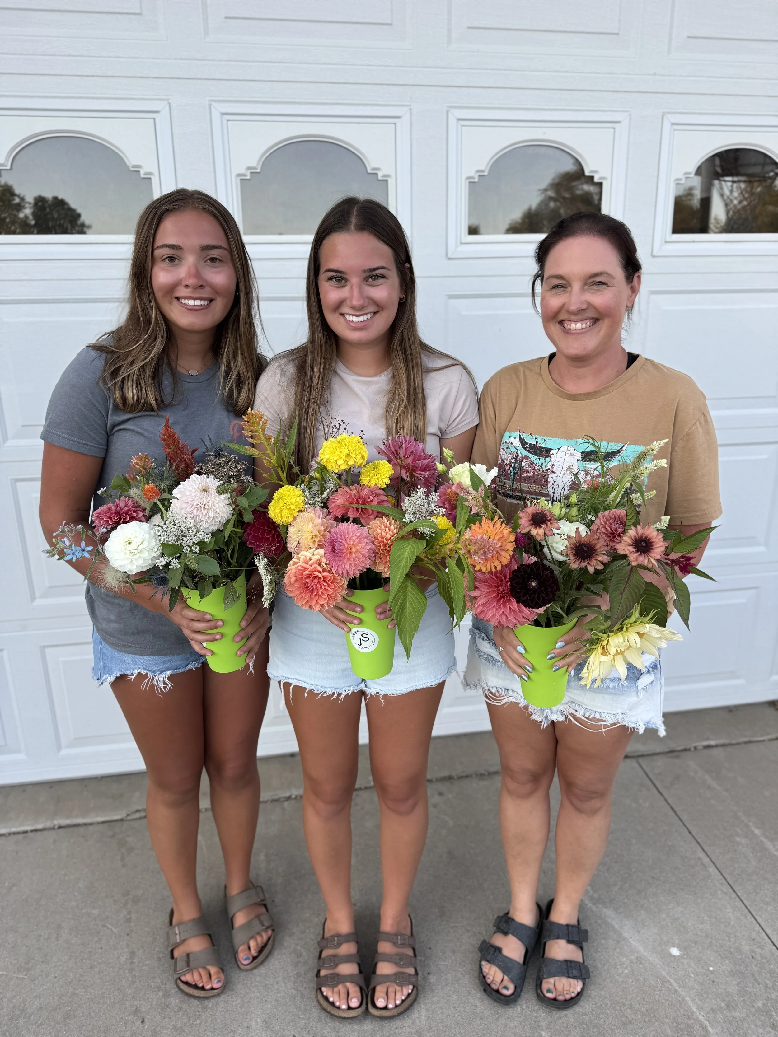 Three women standing outdoors in front of a white garage door, each holding colorful flower arrangements in green pots, smiling at the camera.