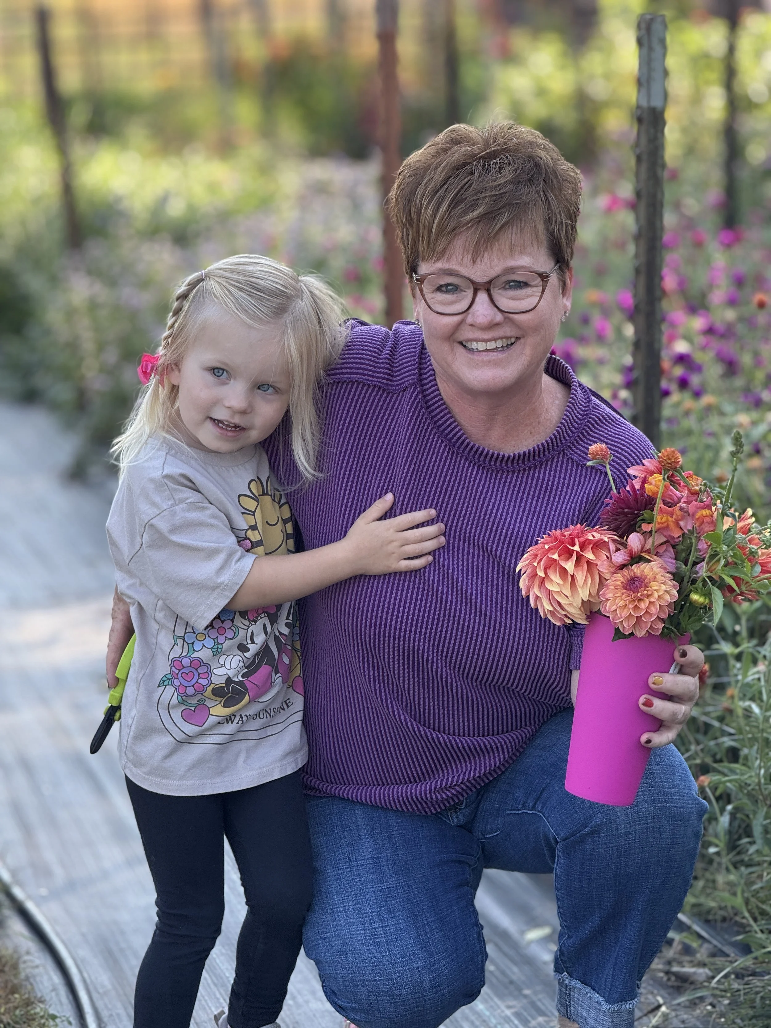 A smiling woman with short hair and glasses holding a pink vase with orange and purple flowers, standing next to a young girl with blonde hair and a gray Minnie Mouse t-shirt outdoors in a garden with colorful flowers.