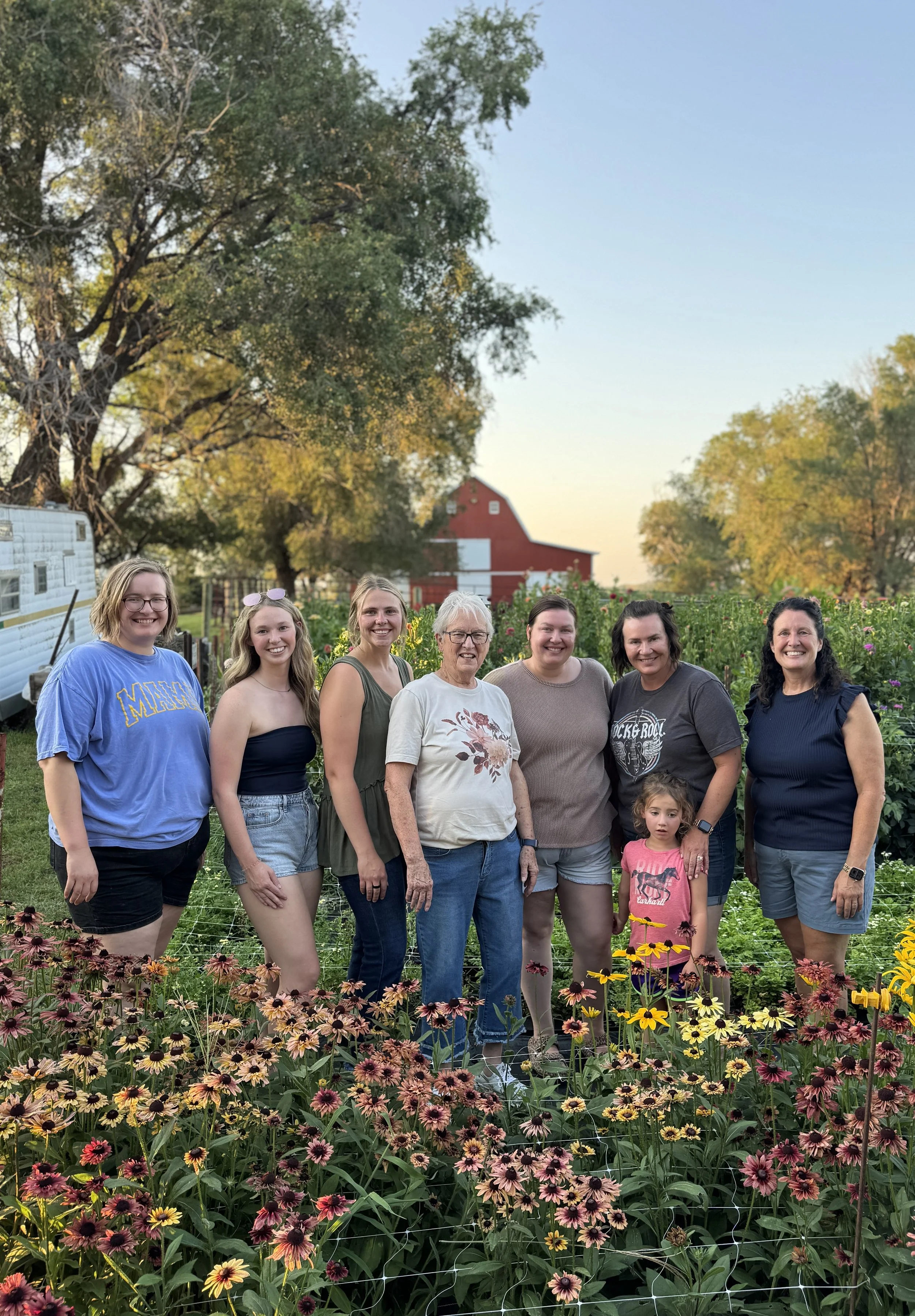 Group of eight women and one young girl standing together in a garden with colorful flowers, trees, and a red barn in the background during sunset.