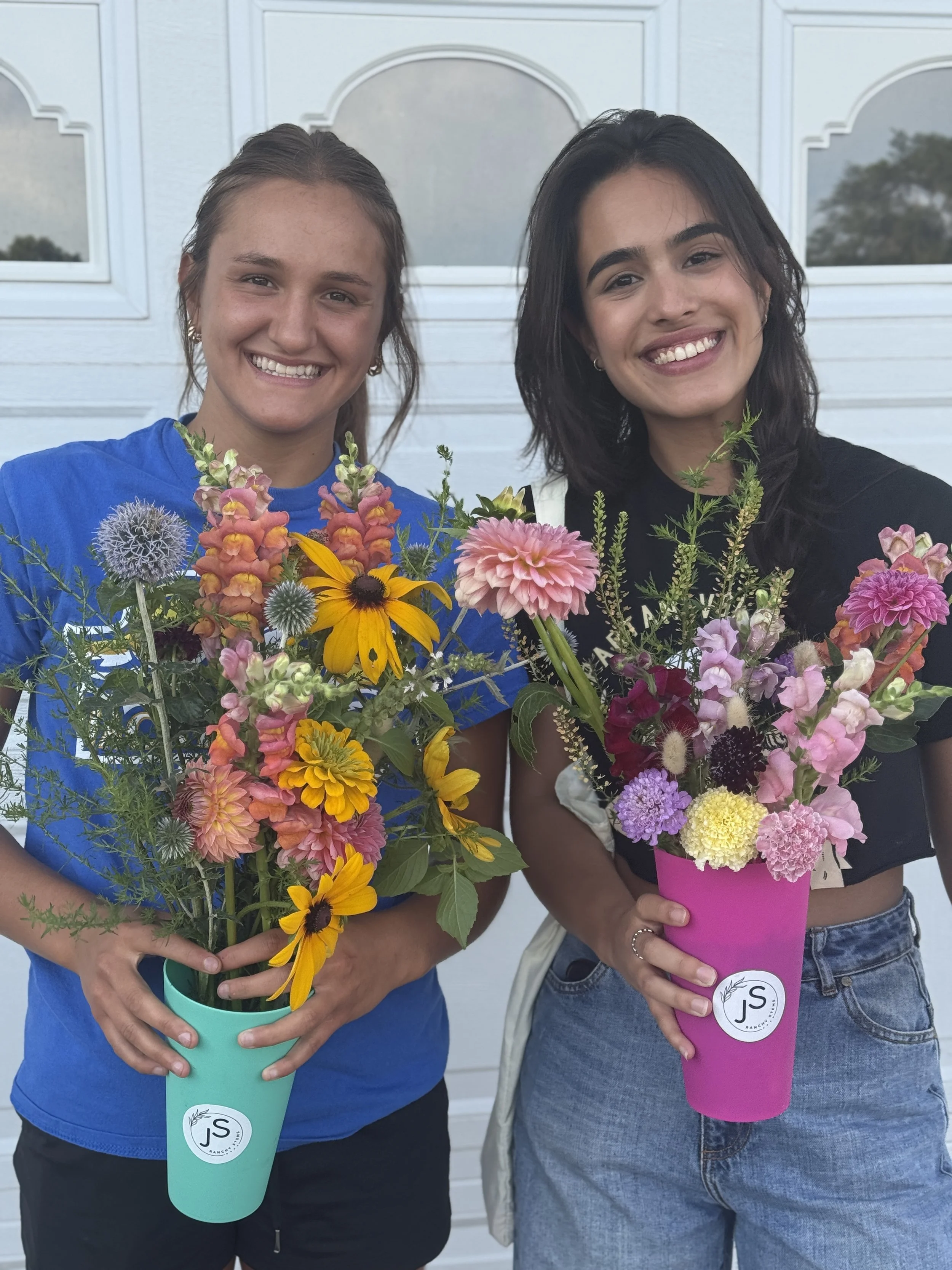 Two women smiling and holding colorful flower bouquets in teal and pink cups outside in front of a white garage door.