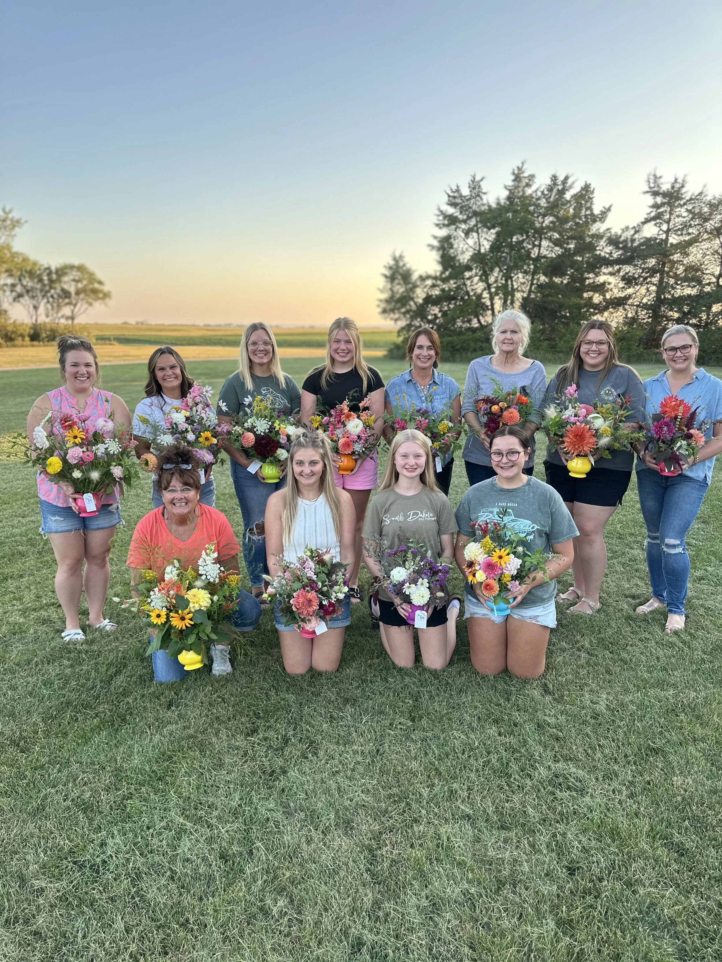 A group of 14 women and girls outdoors on a grassy field in the evening, holding colorful flower bouquets, smiling for a photo with a sunset and trees in the background.