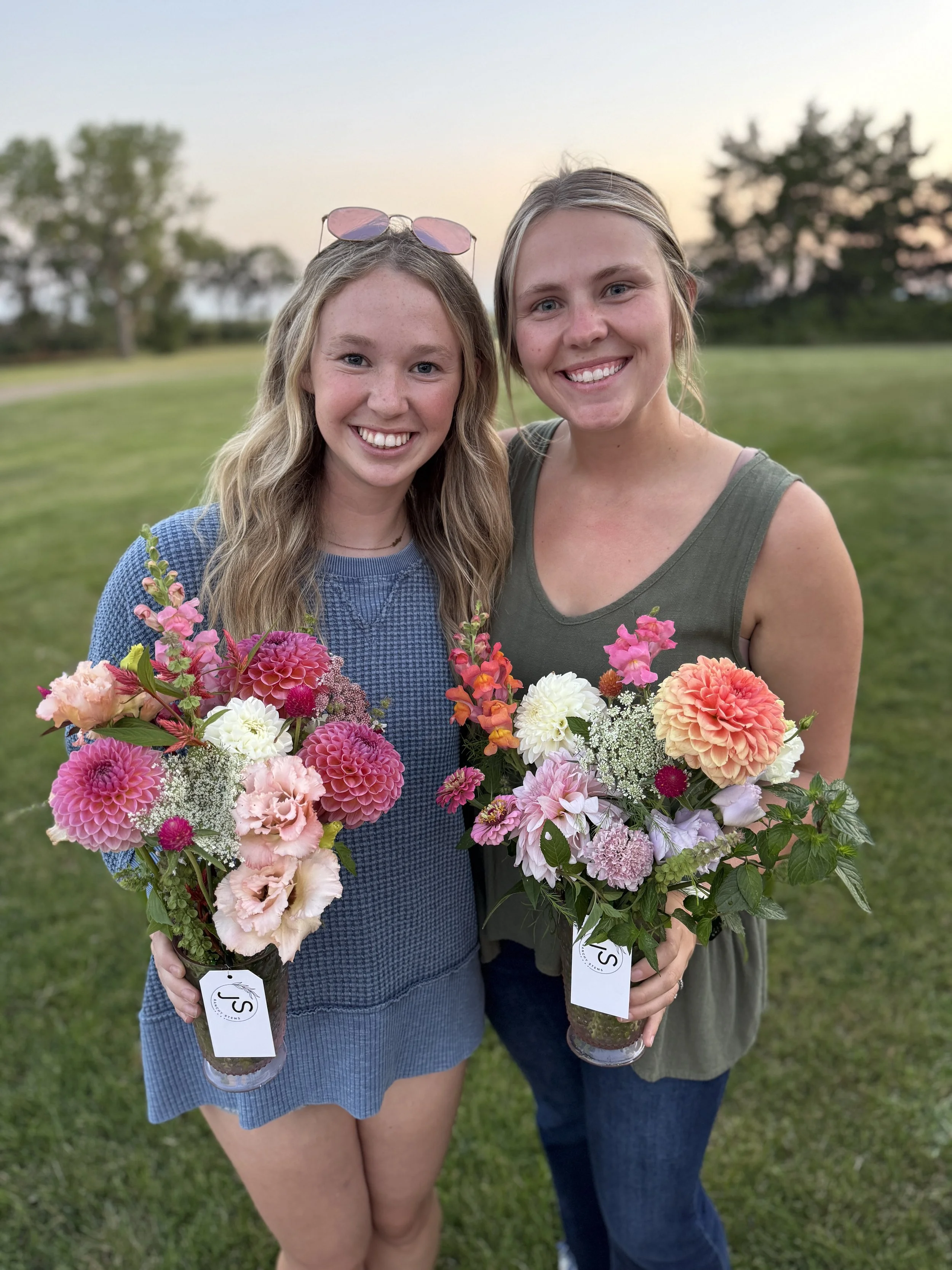 Two young women smiling outdoors at sunset, each holding a bouquet of pink, white, and peach flowers.