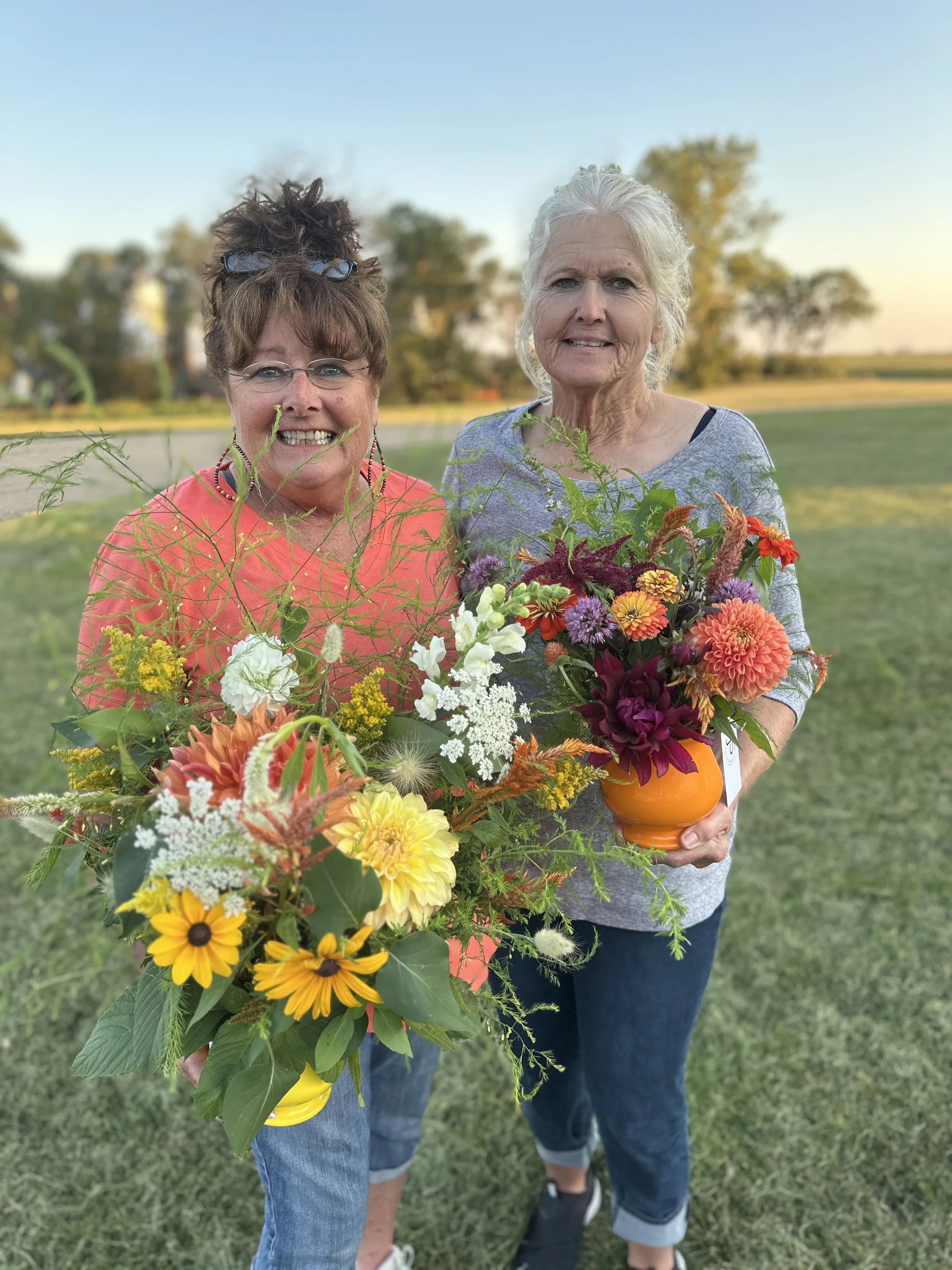 Two women holding colorful flower bouquets outdoors with trees and grass in the background.