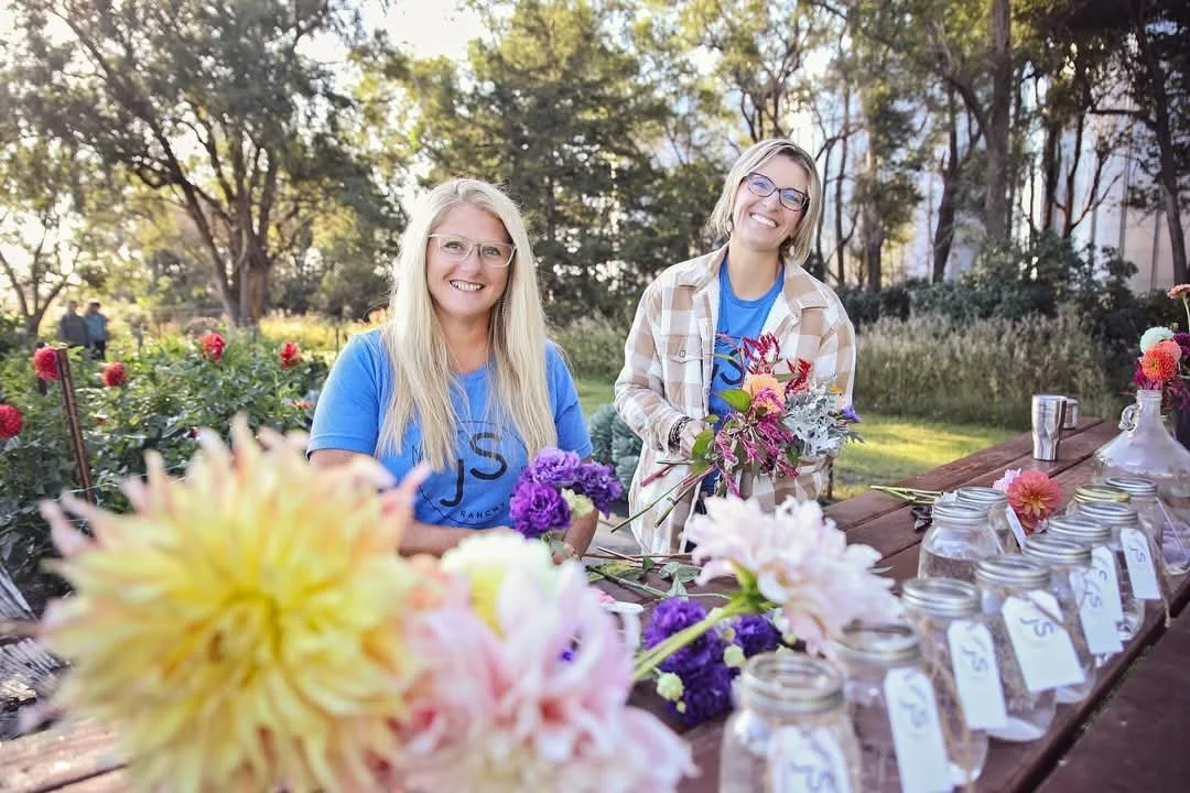 Two women with glasses and long hair standing behind a table with flowers and jars in an outdoor garden park during the daytime.