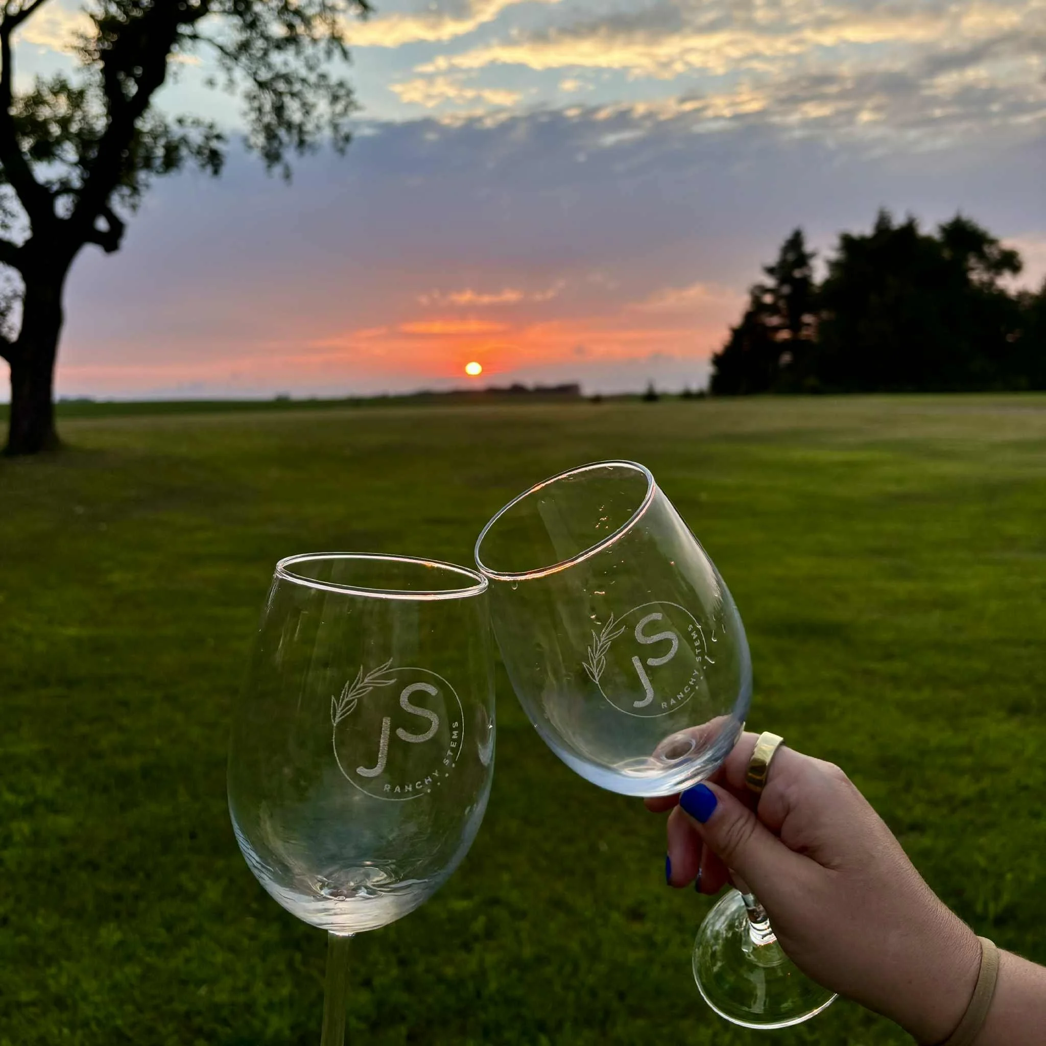 Two wine glasses with 'JS Ranch Systems' logo being held up during a sunset in a grassy field with trees in the background.