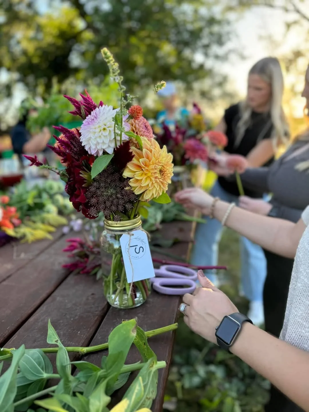 A group of people creating flower arrangements outdoors at a long wooden table, with a focus on a bouquet of assorted flowers in a glass jar, featuring dahlias, celosia, and other colorful blooms.