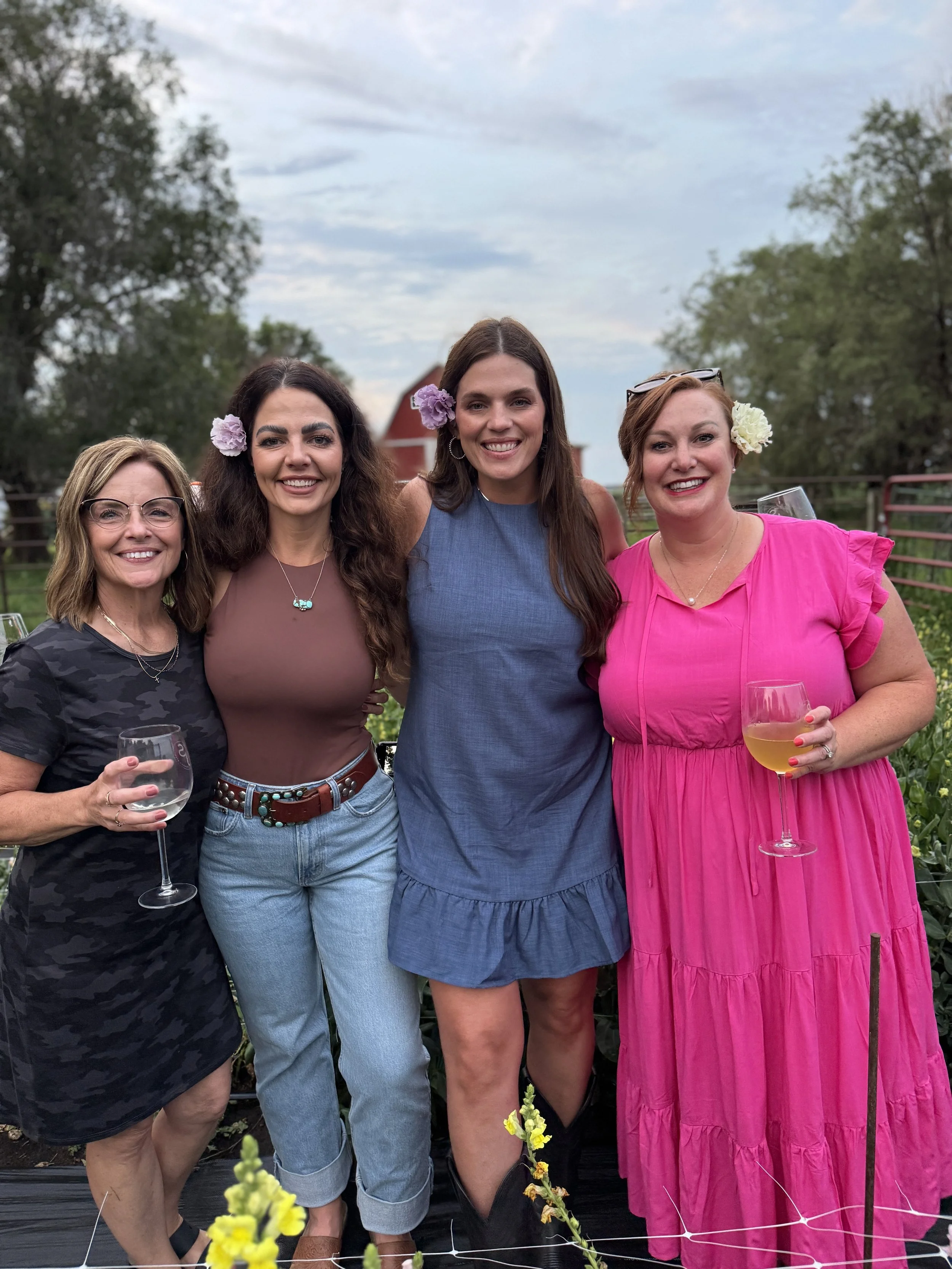 Four women standing outdoors in a garden, smiling, holding wine glasses, wearing colorful dresses, with a farm building and trees in the background.