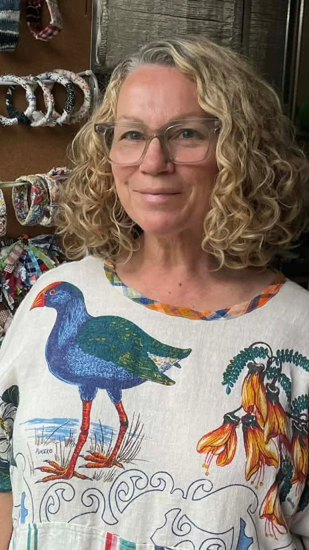Catherine stands in front of her textile jewellery wearing a dress made out of a linen tablecloth. An image of a water hen is on her right shoulder.