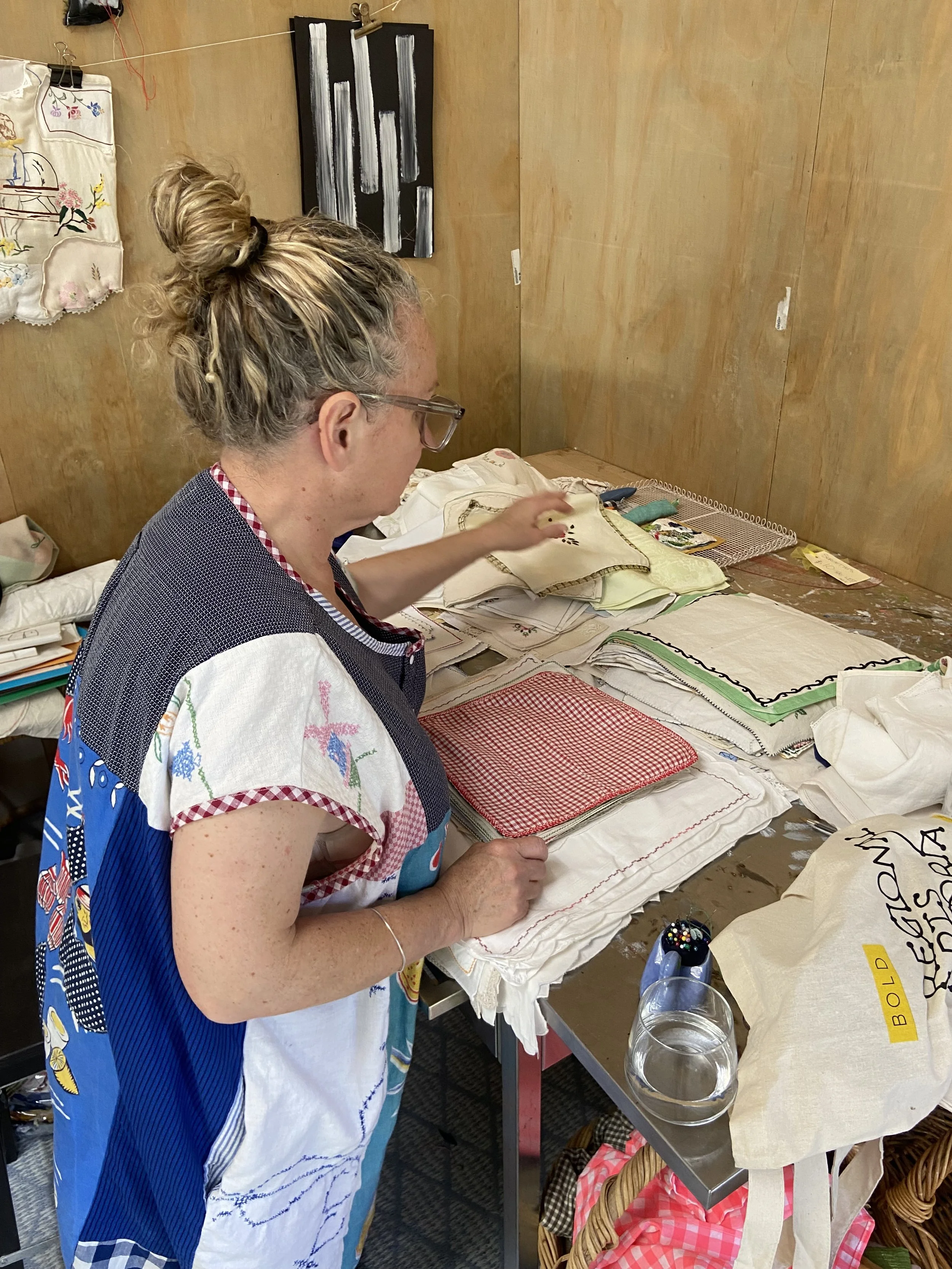 Woman with blond hair in a messy bun, wearing glasses and a colourful dress, working on embroidered textiles on a table covered with fabric and embroidery pieces, in a sewing studio.