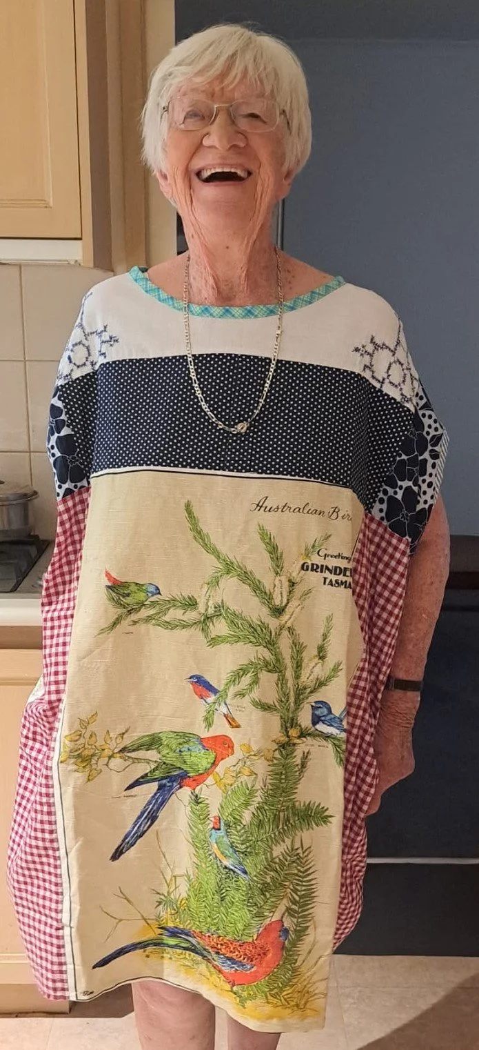 A woman with short white hair and glasses, smiling in a kitchen, wearing a colourful dress featuring various birds and green foliage.