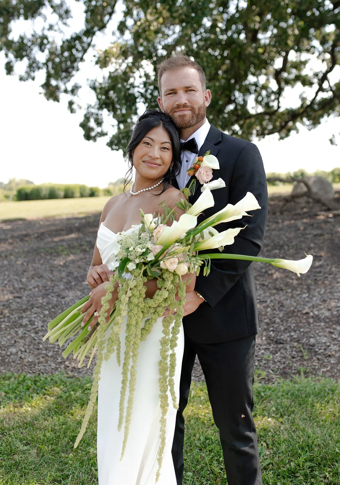 A newlywed couple standing outdoors, the woman in a white wedding dress holding a large bouquet of white calla lilies and greenery, the man in a black tuxedo with a boutonniere, both smiling, with a large tree and open field in the background.