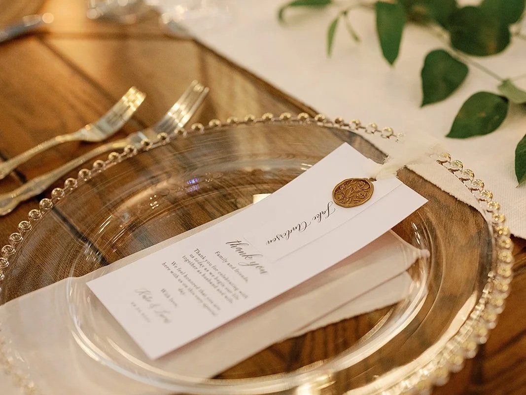A decorative, ornate glass plate with a beaded edge, holding a folded napkin, a printed menu, and a white tag with a gold wax seal, set on a wooden table with silverware and greenery nearby.