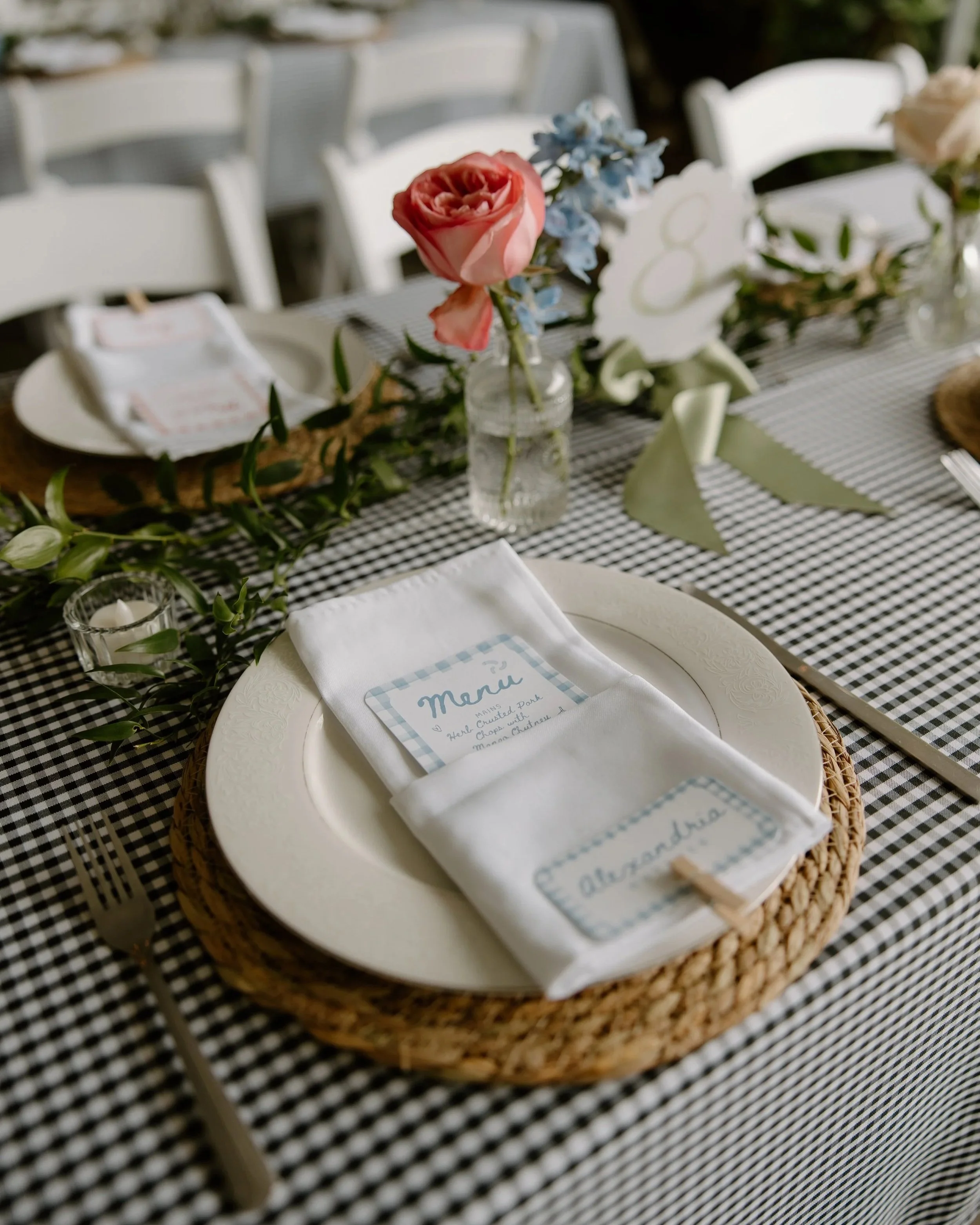 A table setting with a white plate on a woven placemat, a folded white napkin with a menu card and a name tag, surrounded by a garland and small candle holder, with a flower vase containing pink and blue flowers as a centerpiece.