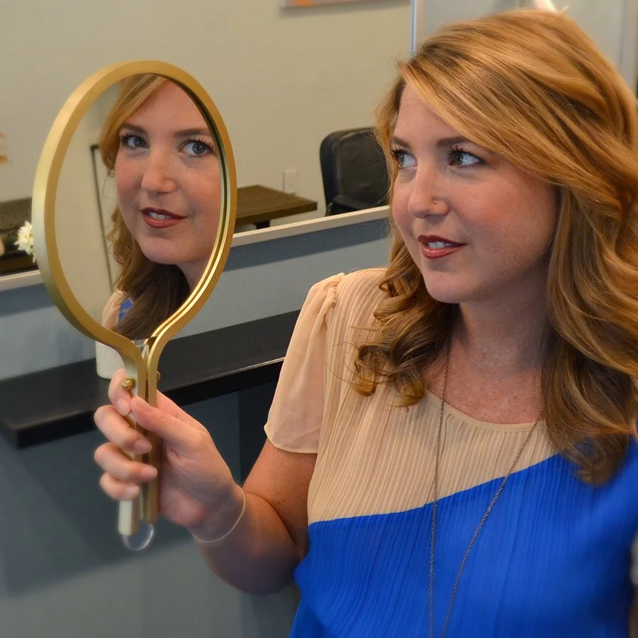A woman with red hair holds a mirror and looks at her reflection.