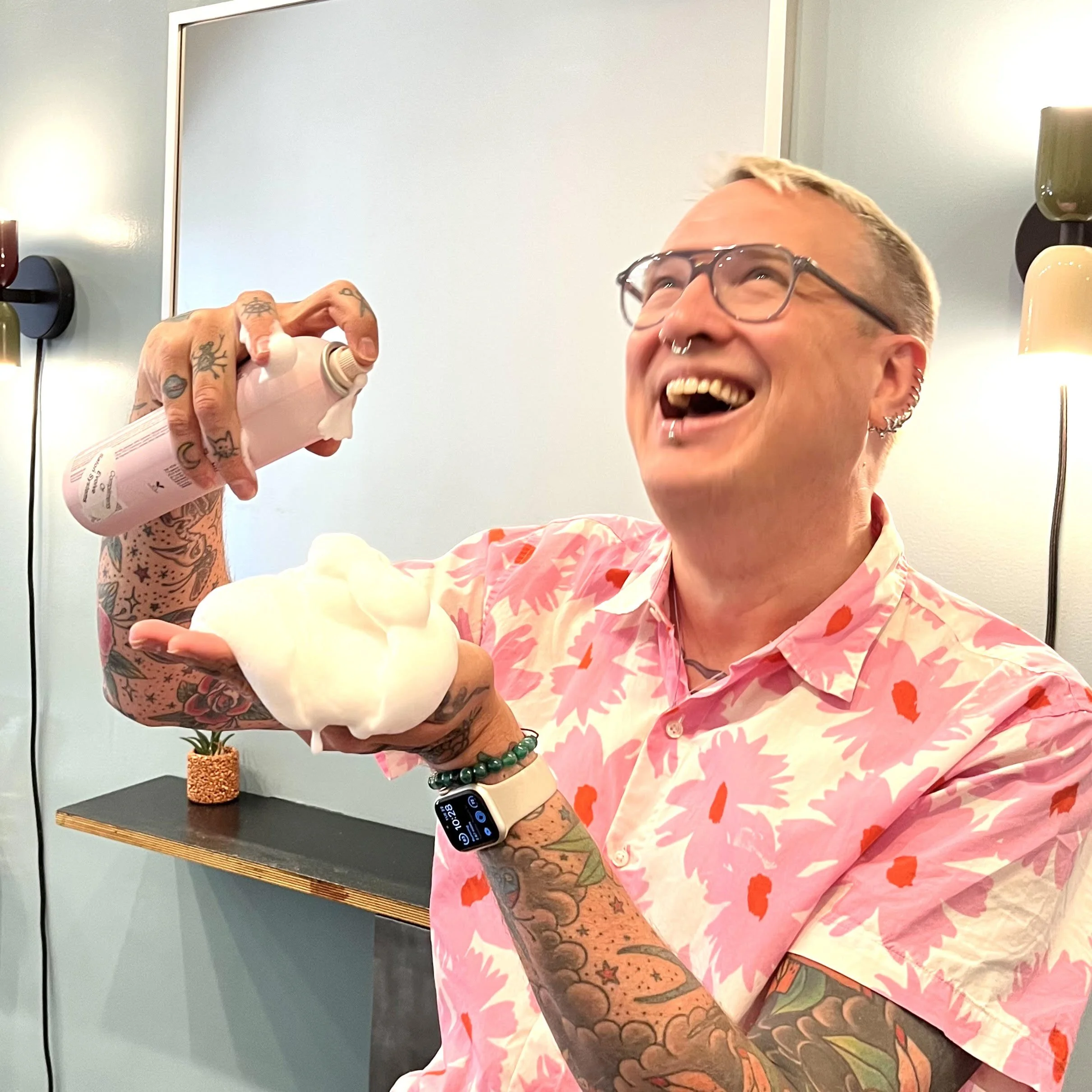 A man with tattoos, glasses, a septum piercing, and multiple earrings, wearing a pink shirt with orange and red floral patterns, smiling and holding a foam doll in one hand and a white spray can in the other.