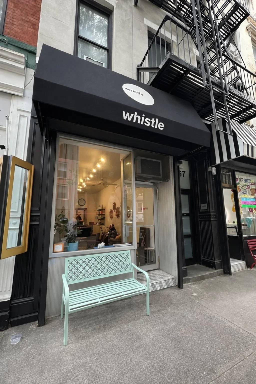 Exterior of a storefront with a black awning labeled 'whistle' in lowercase white letters, a large front window revealing the interior, and a light green bench outside. The building has a fire escape on the upper floors and neighboring storefronts with striped awnings.