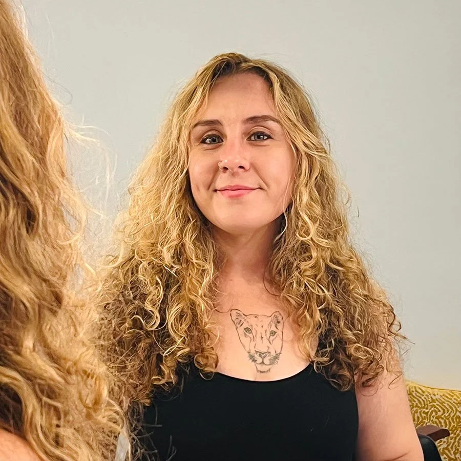 A woman with curly blonde hair wearing a black top, smiling in front of a light-colored wall. She has a tiger face tattoo on her chest.