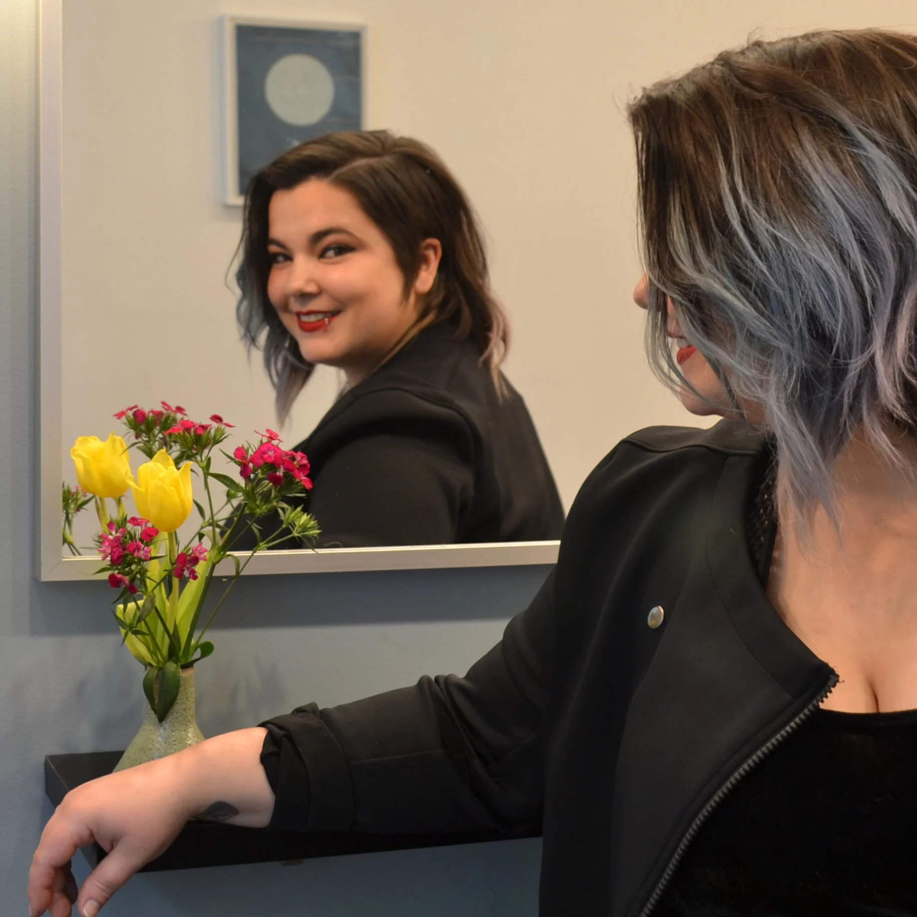 A woman with shoulder-length gray hair and a black leather jacket looking at a mirror, smiling. In the mirror, a woman with short dark hair and red lipstick is smiling back. A vase with yellow tulips and pink flowers is on the counter.