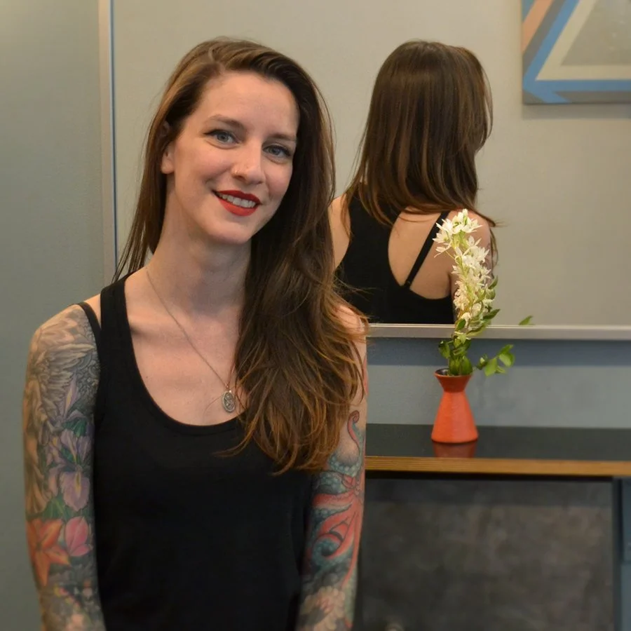 A woman with long brown hair, red lipstick, and tattoos on her arms, smiling and wearing a black tank top, sitting in front of a mirror with a vase of white flowers on a table behind her.