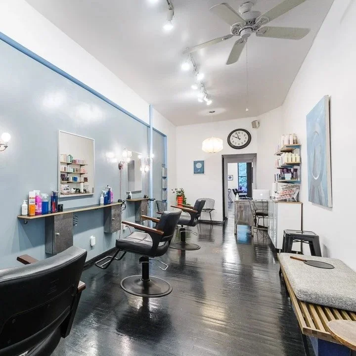 Empty hair salon with styling stations, mirrors, chairs, and shelves with hair products, bright lighting, hardwood floors, and a large wall clock.