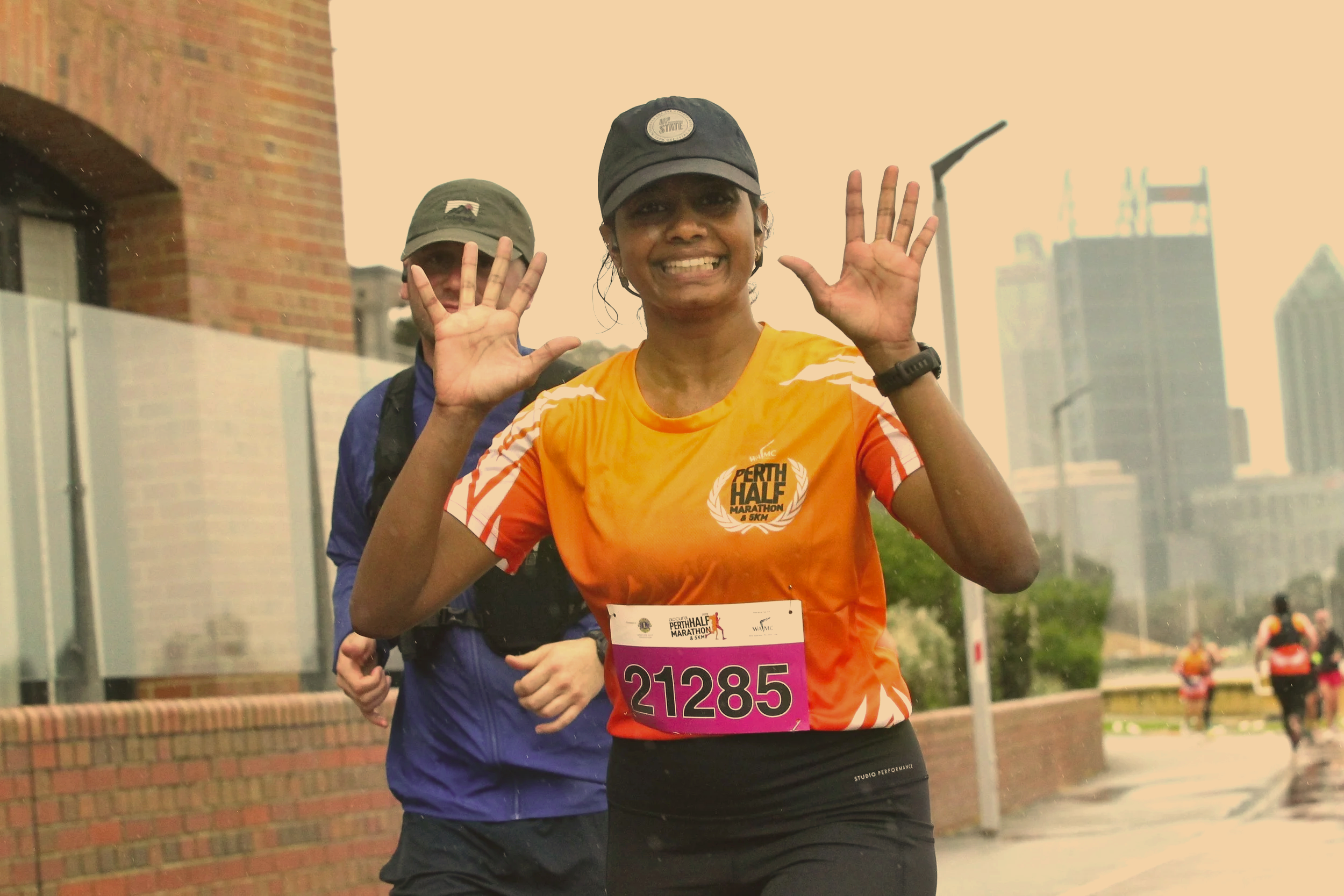 A woman in an orange running shirt smiling and waving, participating in a marathon, with a man in a blue jacket and gray hat behind her, both running outdoors on a city street with buildings and trees.