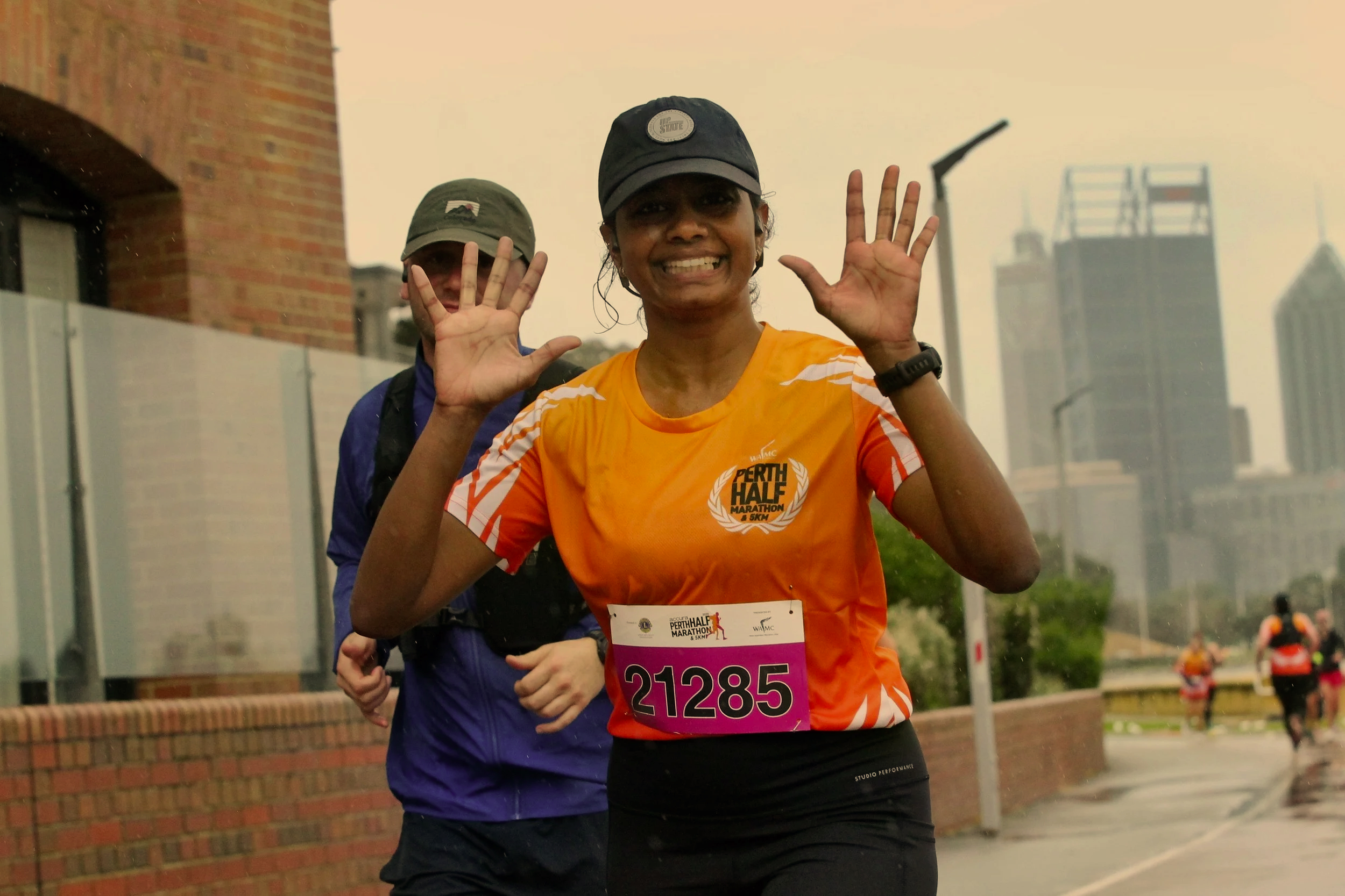 A woman running in a marathon wearing an orange shirt, black shorts, a black cap, and a bib number 21285. She is smiling and holding her hands up in a peace sign. A man is running behind her wearing a green cap and a blue jacket. The background features a brick building and some greenery, with other runners visible in the distance.