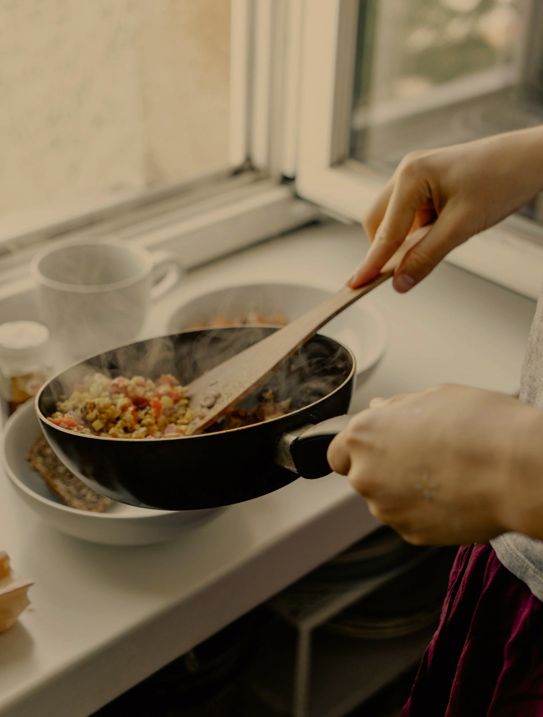A person is cooking on a stove with a black frying pan, stirring food with a wooden spatula. The stove has a white surface, and there are cups and dishes in the background near a window.